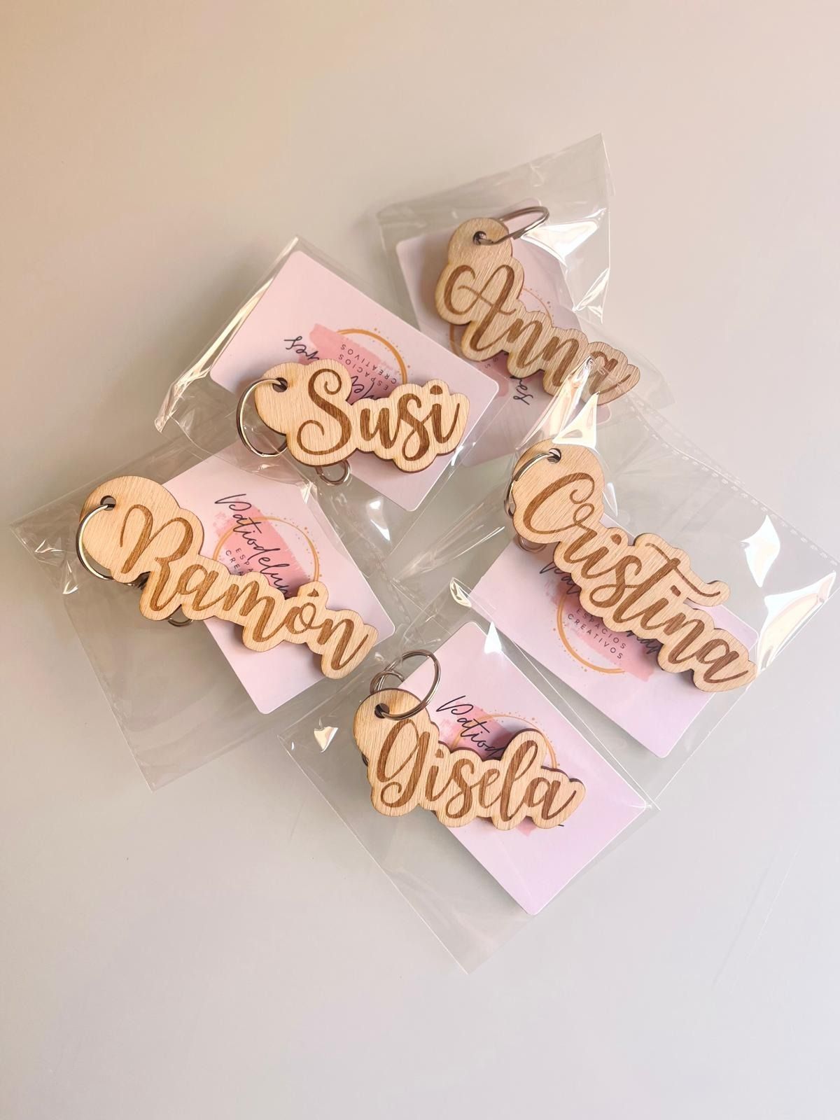 Cinco llaveros de madera personalizados con el nombre del personaje, cada uno en un embalaje individual de plástico transparente con una tarjeta de respaldo de color rosa claro.