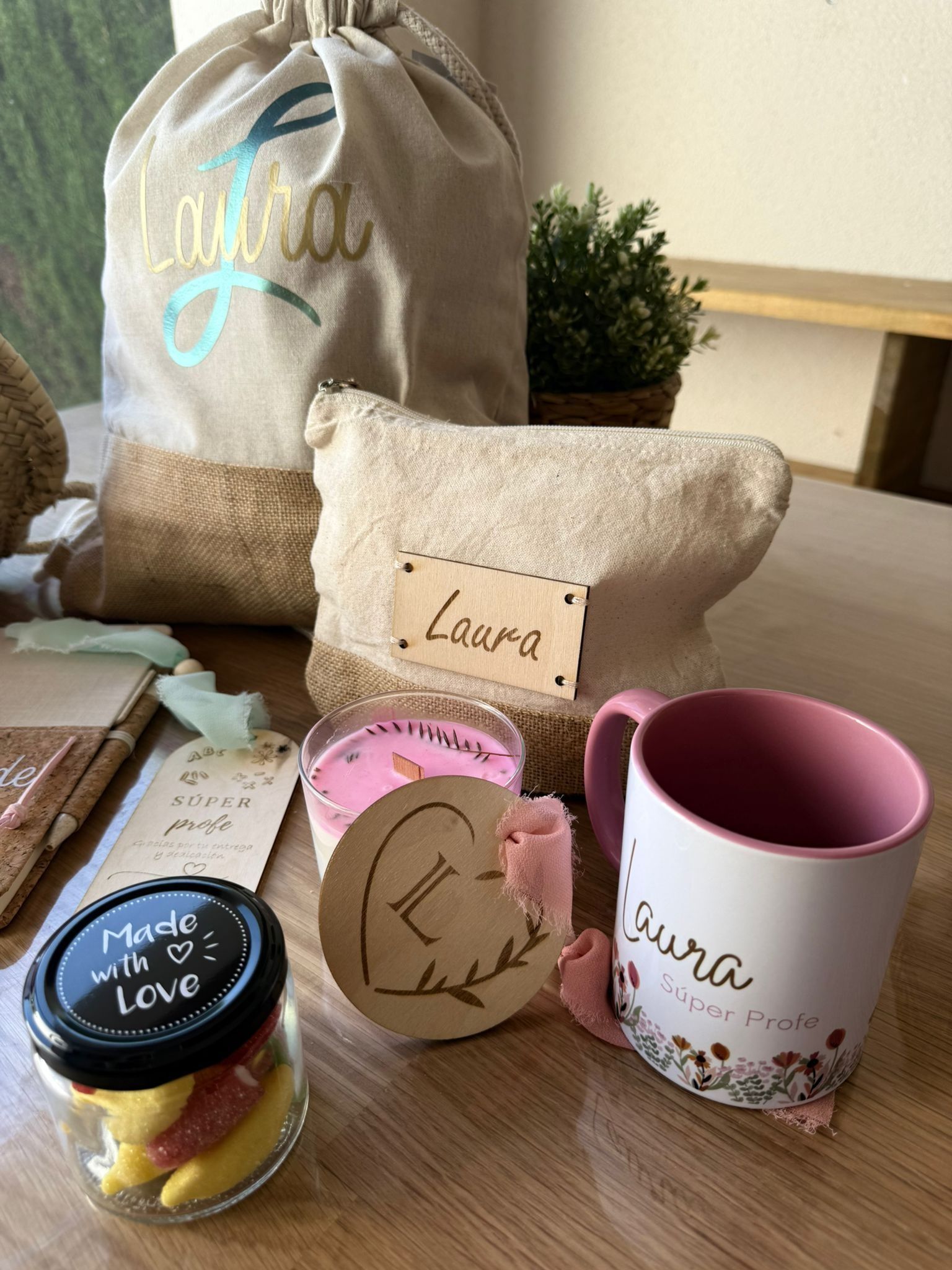 Un set de regalo para Luara que incluye una bolsa personalizada, una taza, una vela, una pequeña planta y un tarro de caramelos de goma sobre una mesa de madera.