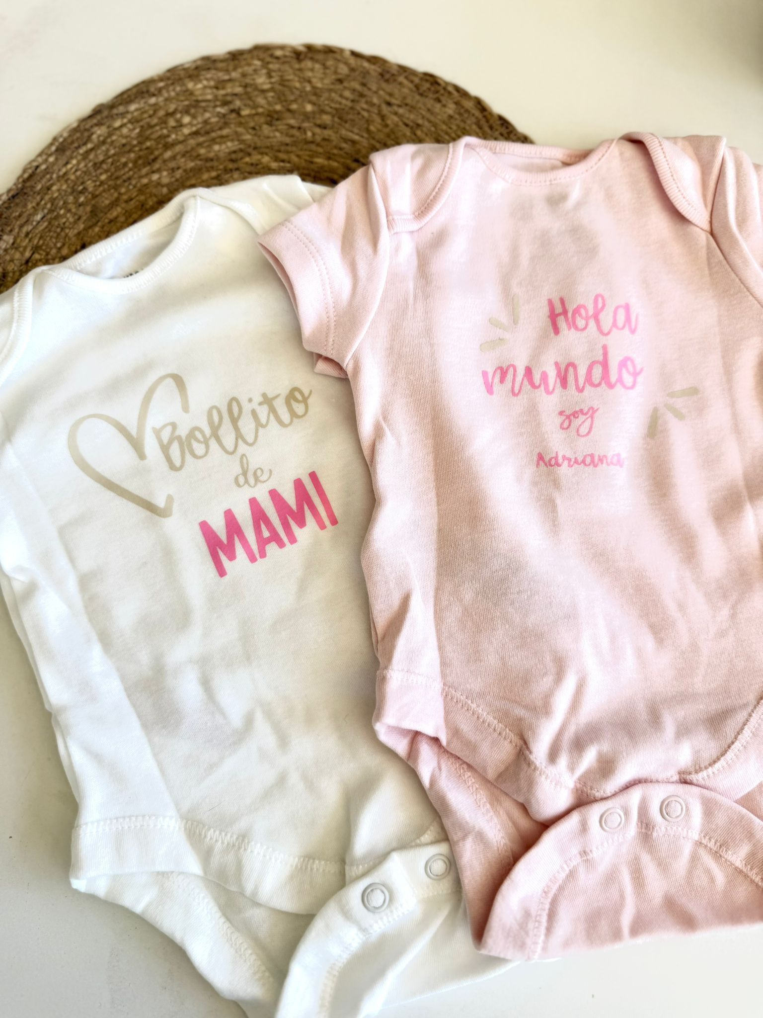 Dos bodys de bebé yacen sobre una superficie tejida. El blanco dice 