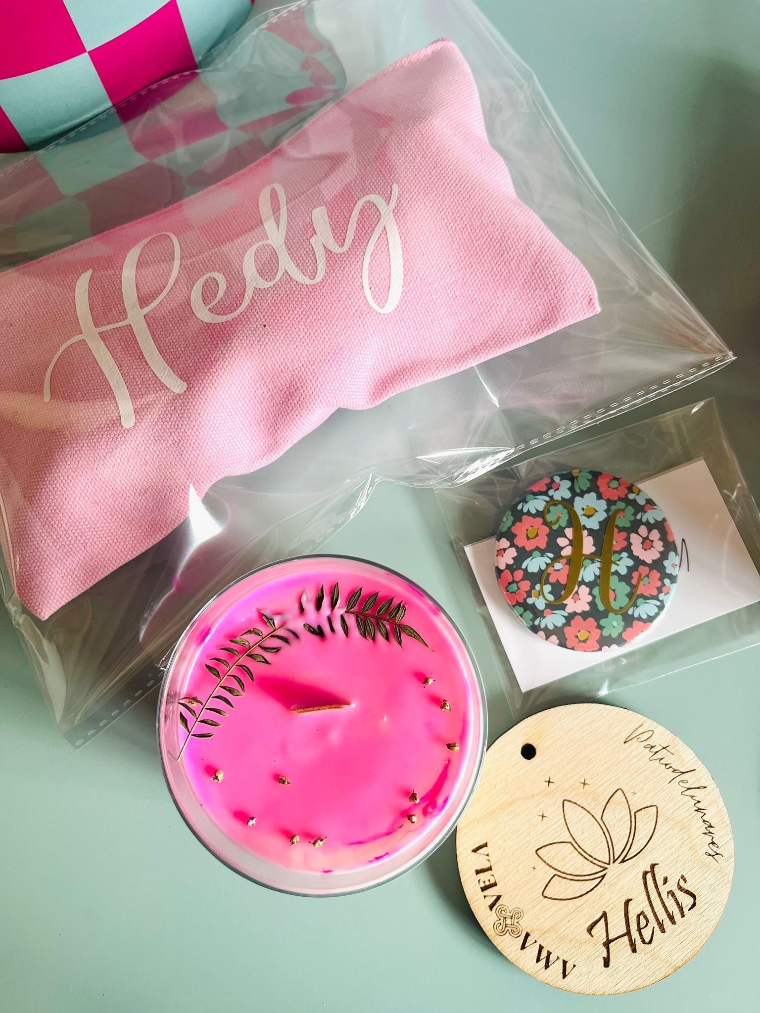 Una bolsa de tela rosa, una vela rosa, una moneda floral con la letra H y una etiqueta redonda de madera con la palabra 