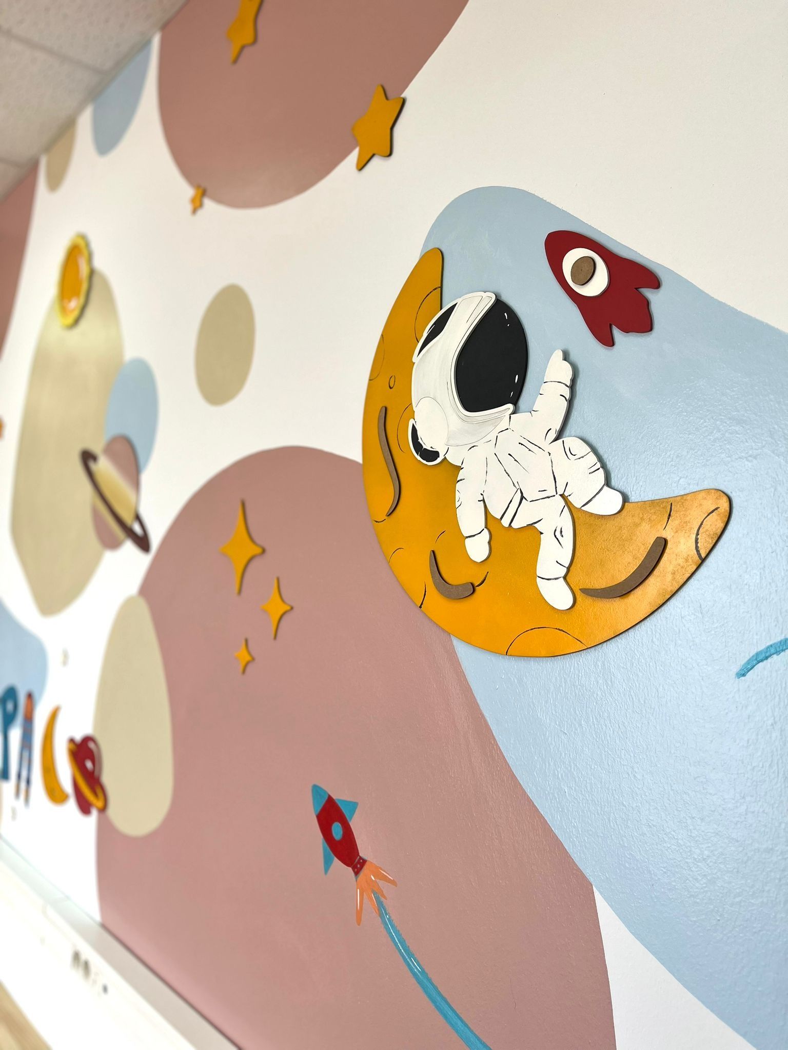 Un mural de pared con temática espacial y un toque de fantasía, que muestra a un astronauta de dibujos animados sentado sobre una luna creciente, planetas y cohetes.