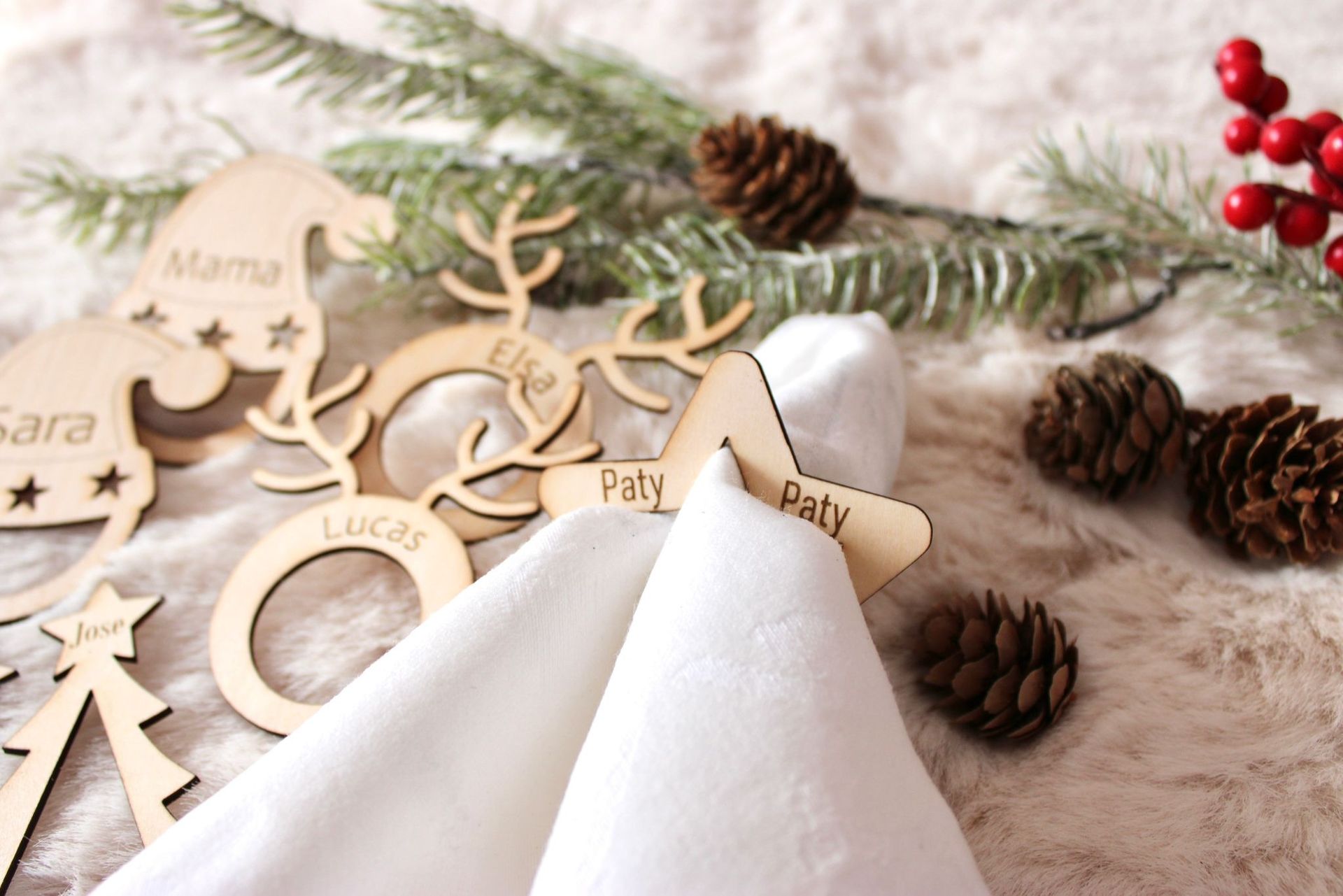 Servilleteros navideños de madera personalizados con nombres, dispuestos sobre un fondo de piel sintética con piñas y ramitas de hoja perenne.