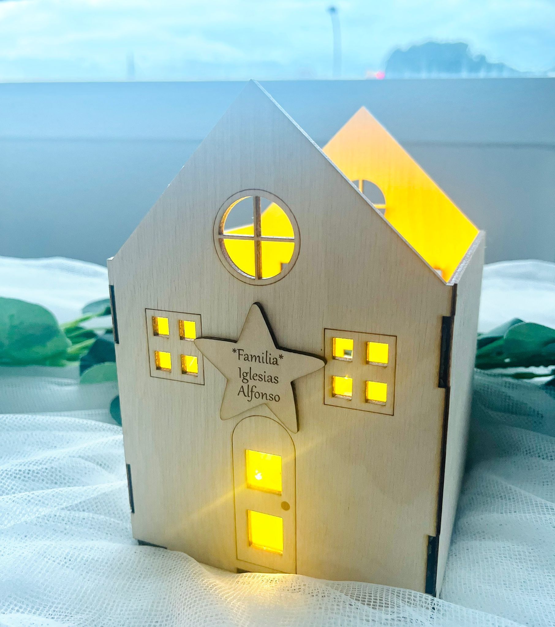 Una linterna de madera cortada con láser con forma de casa y diseño de estrella, que emite una cálida luz amarilla en su interior.