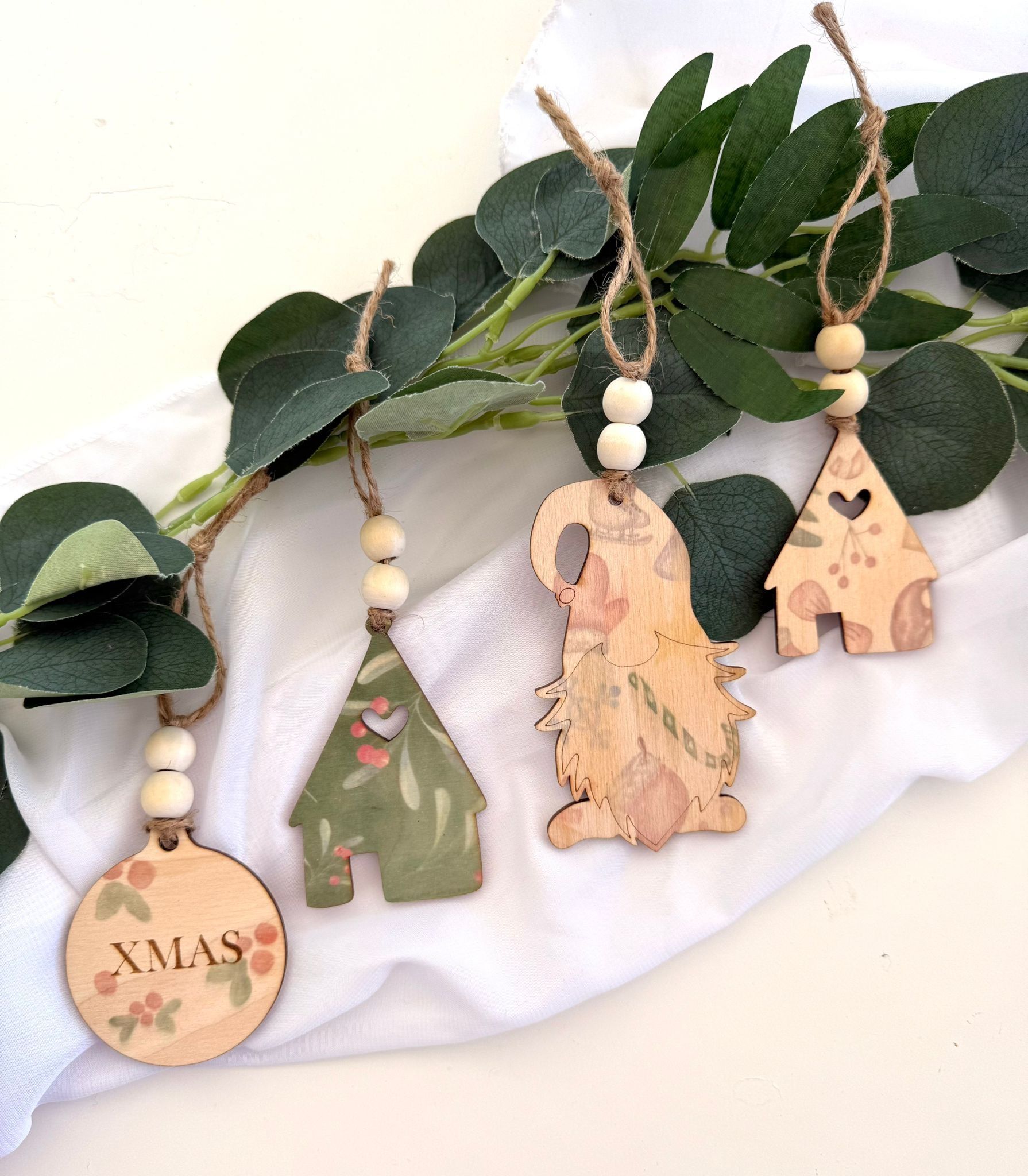Cuatro adornos decorativos de madera, entre ellos una casa y un gnomo, cuelgan de un cordel con cuentas de madera sobre hojas verdes.