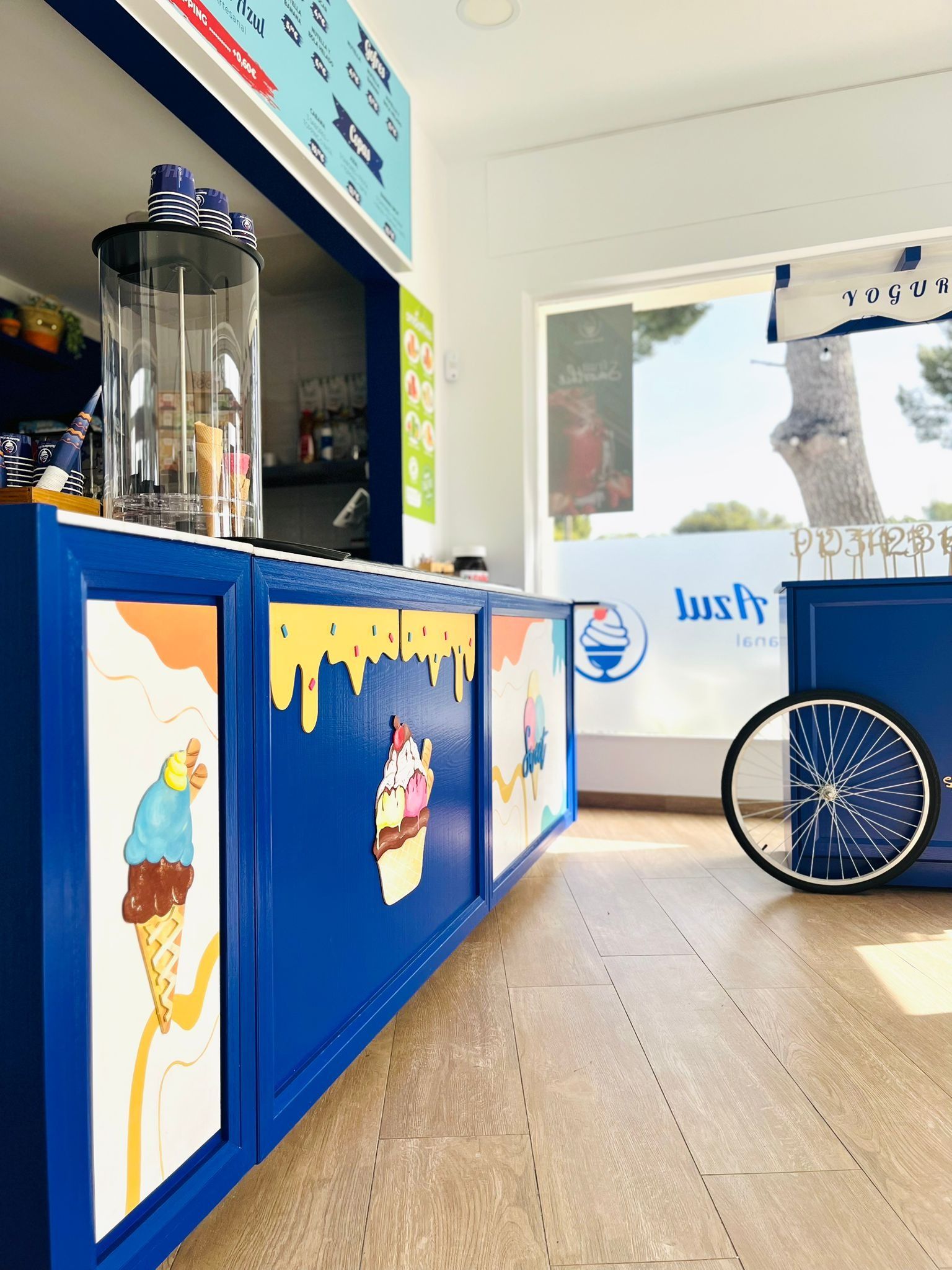 Un mostrador de helados de color azul brillante decorado con conos ilustrados, con una rueda de bicicleta expuesta a la derecha.