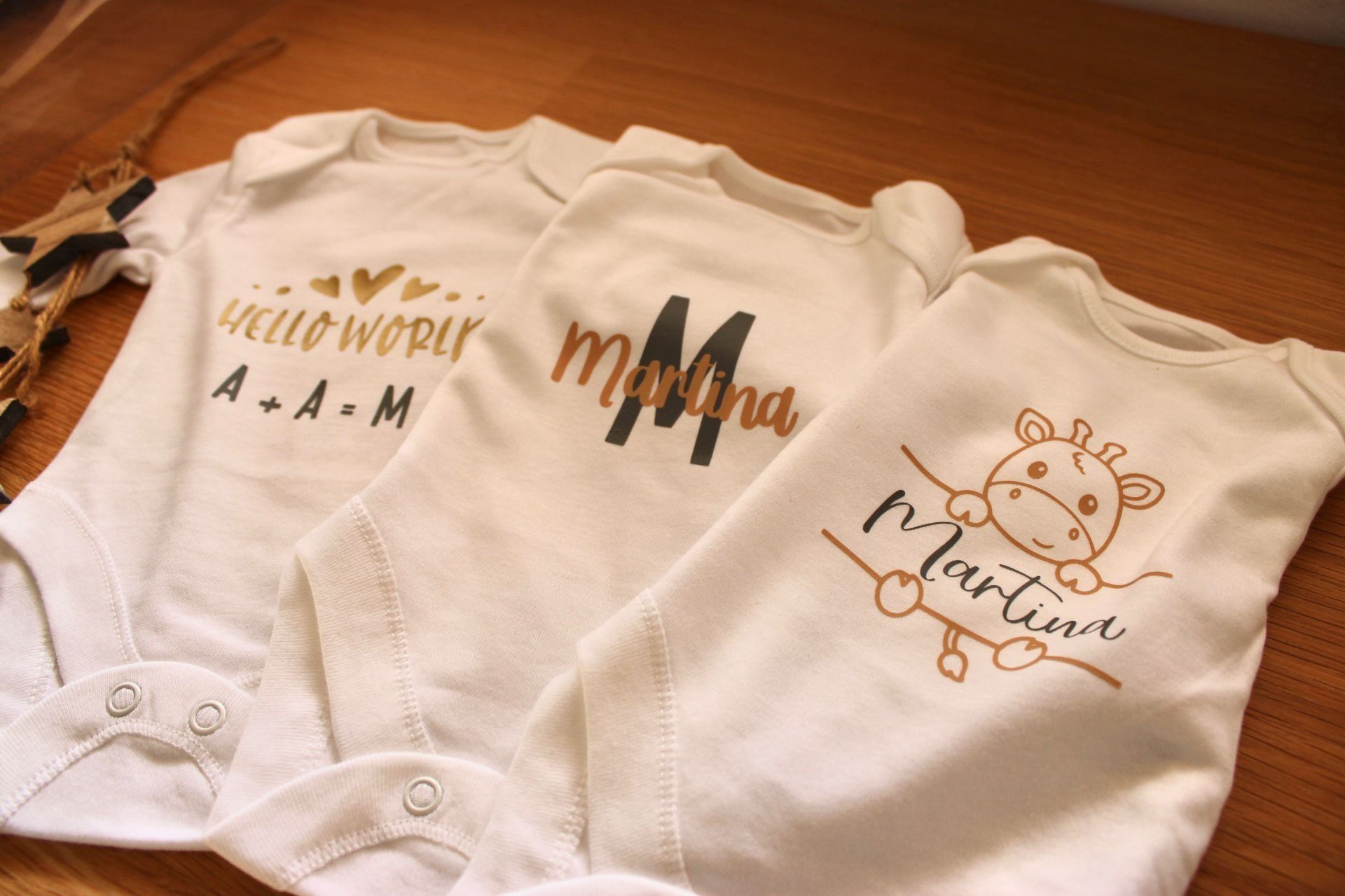 Tres mamelucos blancos para bebé con diseños personalizados estampados en marrón y dorado, sobre una superficie de madera.
