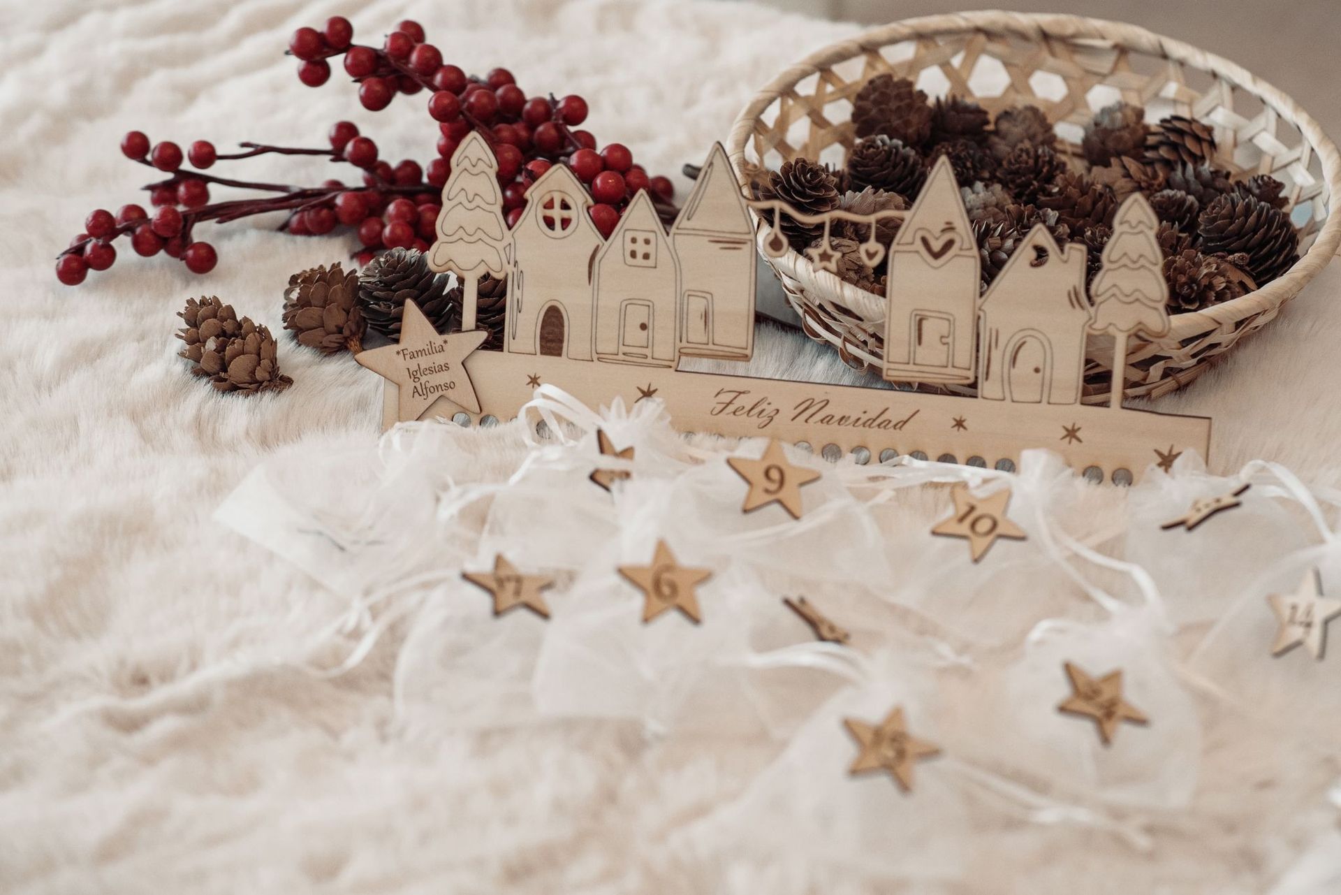 Un adorno navideño de madera con forma de pueblo, con etiquetas numeradas en forma de estrella y pequeñas bolsas, que descansa sobre una superficie blanca y afelpada.