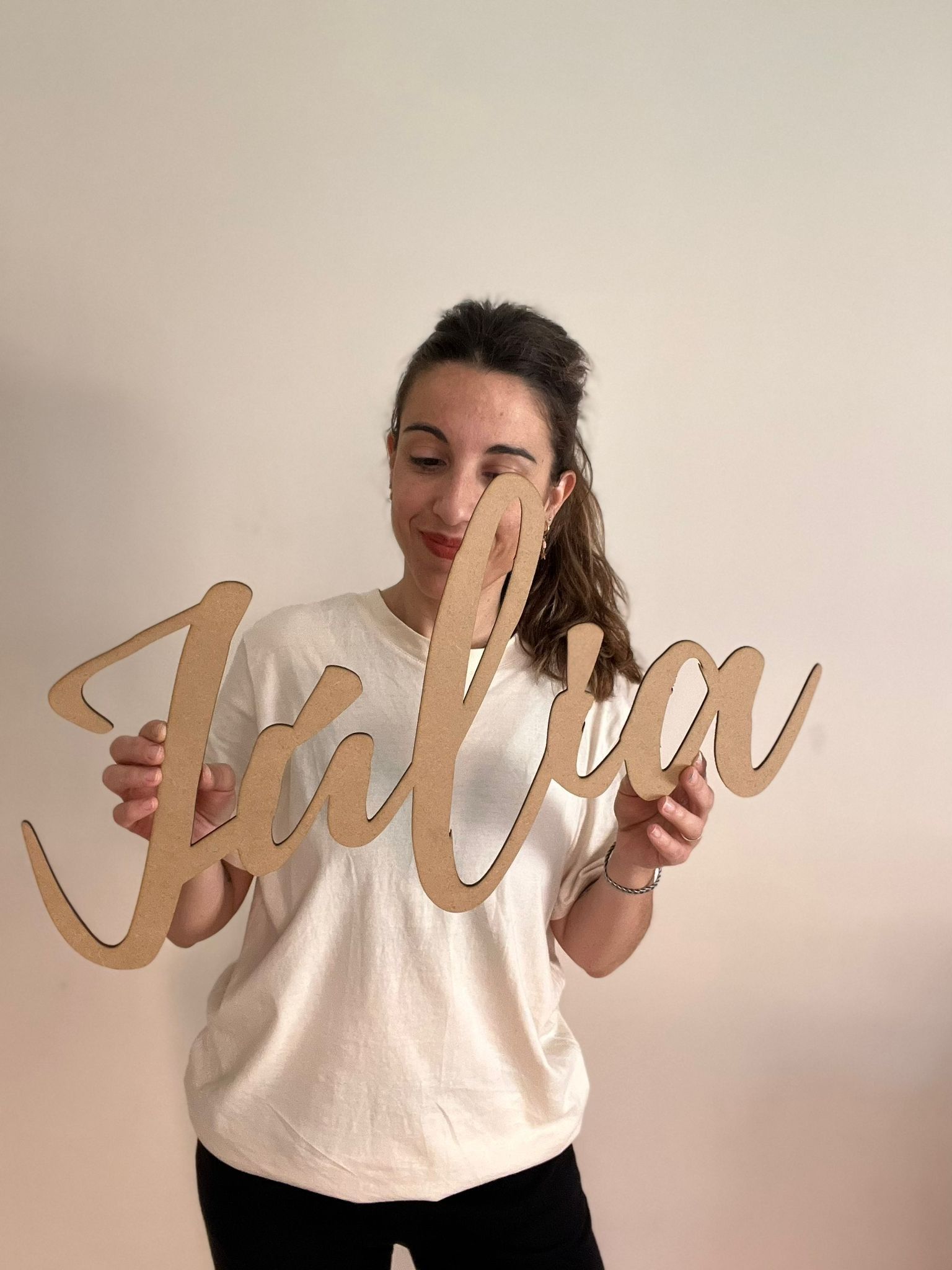 Una persona está de pie junto a una pared blanca, sonriendo mientras sostiene un recorte de madera de color claro con la letra 