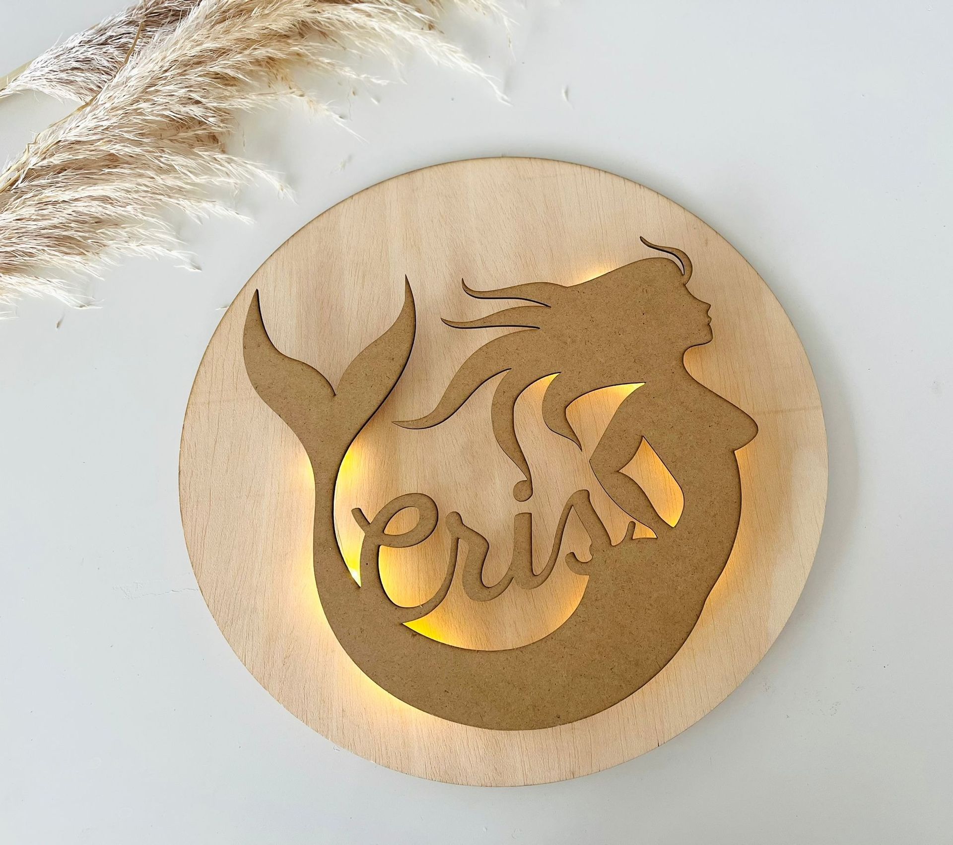 Un adorno de pared circular de madera con la silueta de una sirena recortada, retroiluminada con luz cálida, que lleva el nombre 