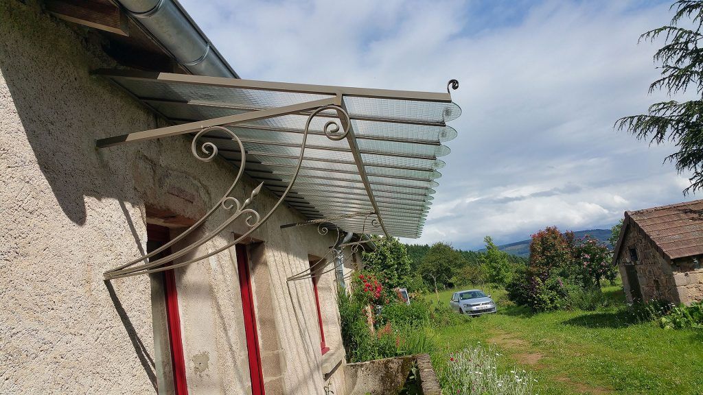 Auvent en verre recouvrant les fenêtres à encadrement rouge d'un bâtiment en pierre, orné de ferronnerie.