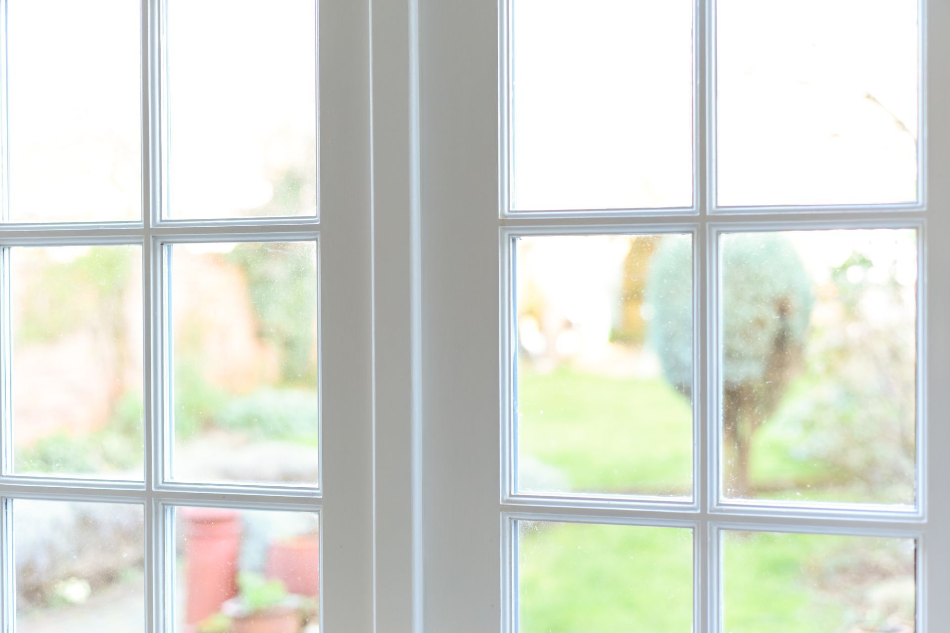 Portes vitrées à cadre blanc et à motif quadrillé, vue légèrement floue sur un jardin verdoyant et des pots rougeâtres.