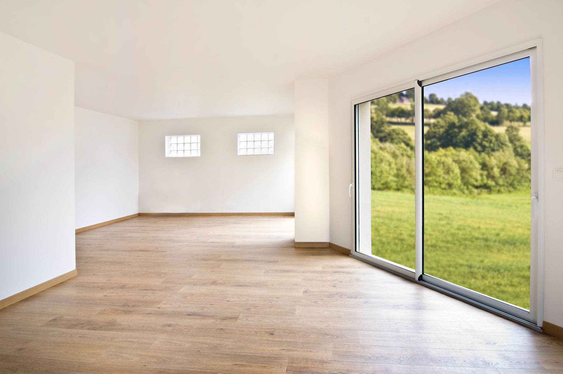 Pièce vide avec parquet, murs blancs et grande fenêtre donnant sur un paysage verdoyant.