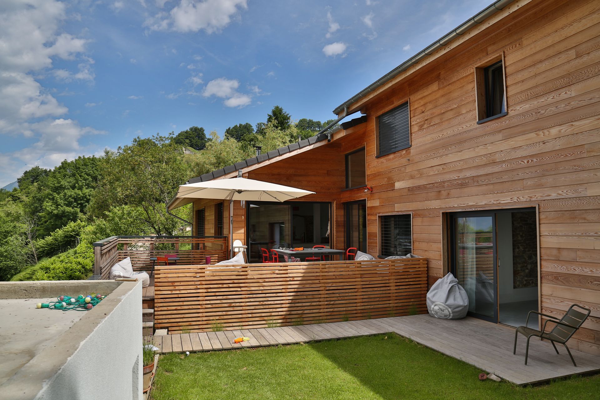 Maison en bois avec terrasse, parasol et jardin, le tout sur fond d'arbres et de ciel bleu.