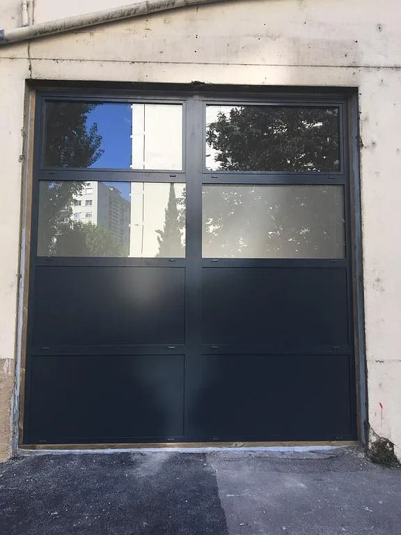 Porte de garage gris foncé avec fenêtres, installée dans un bâtiment en béton clair.