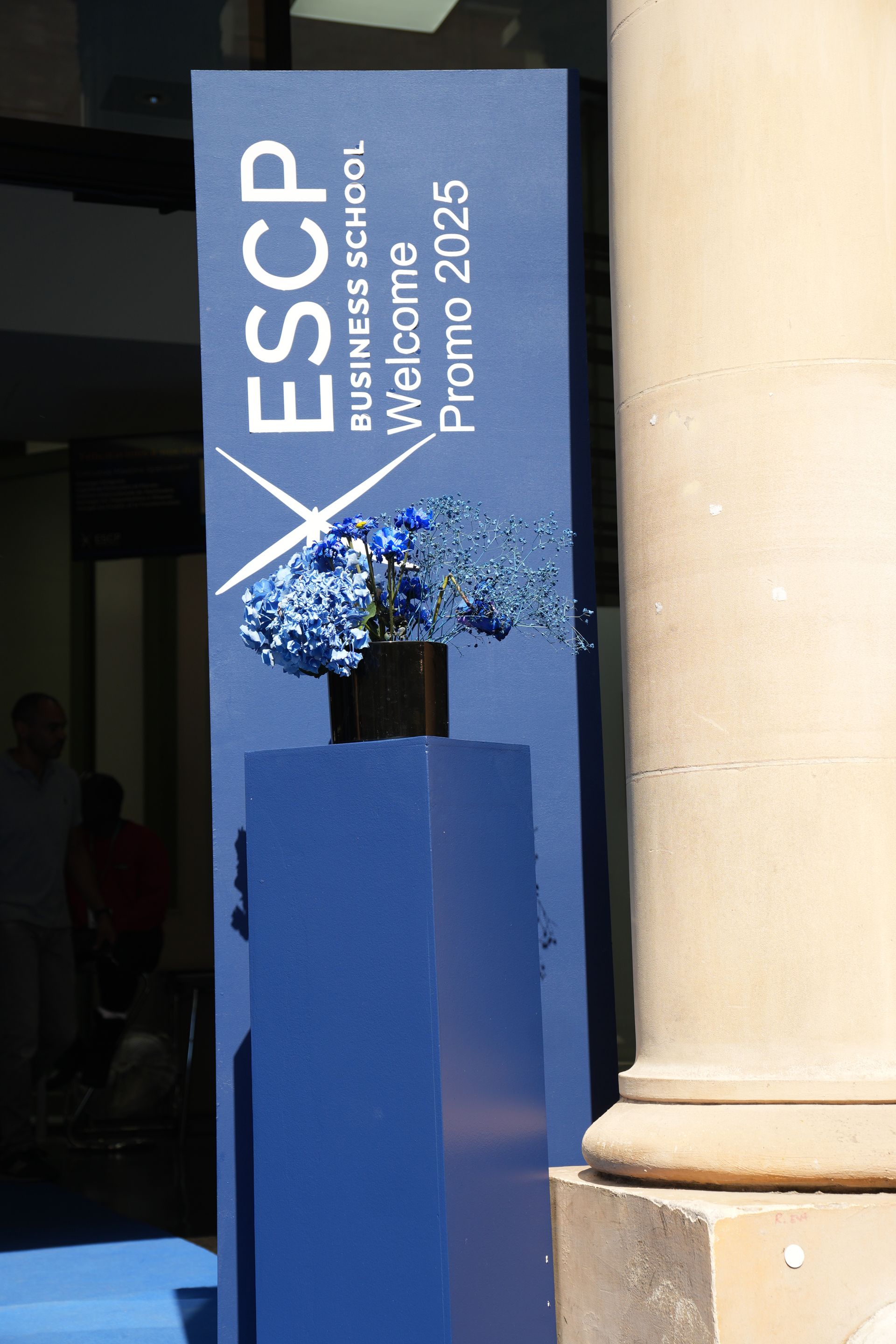 Podium bleu de l'ESCP orné de fleurs et d'une pancarte indiquant « Bienvenue à la Promo 2025 » à côté d'une colonne en pierre.