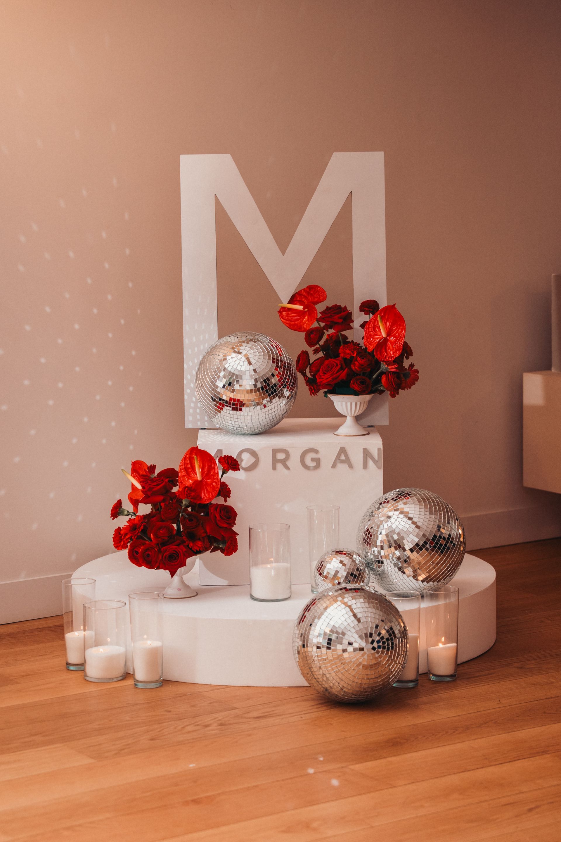 Présentoir décoratif composé d'ornements argentés, de fleurs rouges et de bougies blanches, sur un piédestal blanc avec un grand fond en forme de « M ».