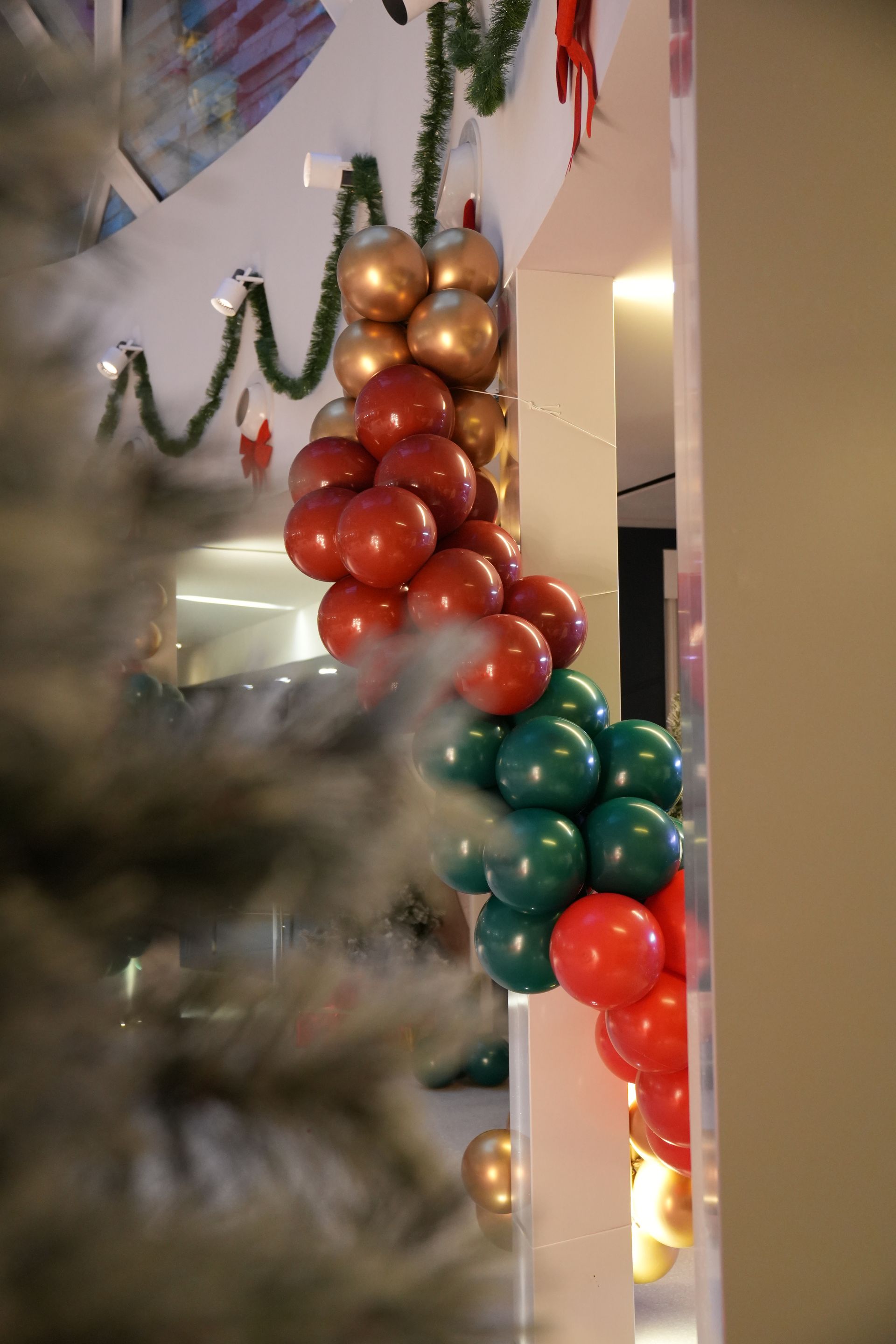 Guirlande de ballons de Noël aux tons métallisés rouge, or et vert, placée à côté d'un sapin de Noël à l'intérieur.