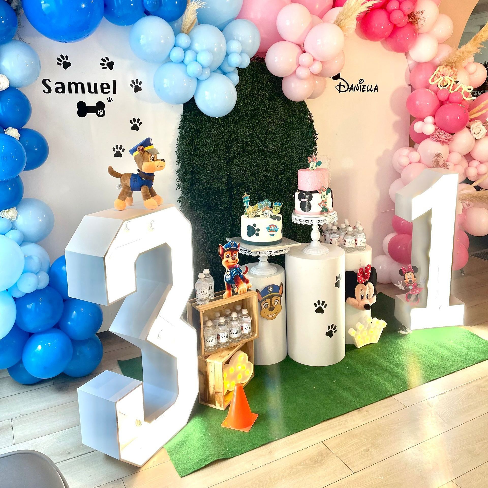 Décoration de fête avec des ballons géants blancs en forme de 3 et 1, une arche de ballons bleus et roses, et des petits gâteaux à l'effigie de personnages sur fond vert.
