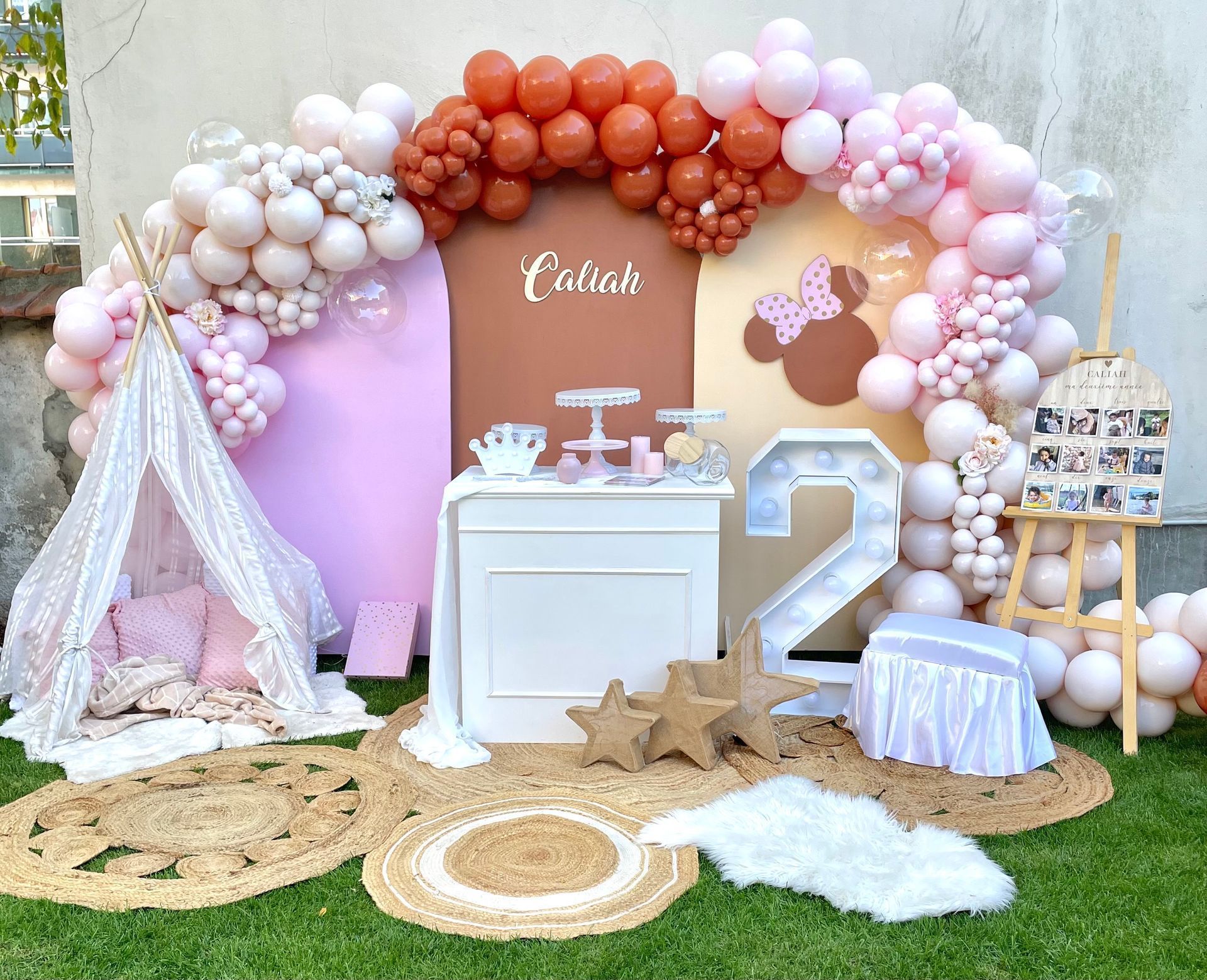 Arche de ballons pastel avec une table de desserts blanche et un grand « 2 » en arrière-plan d'une fête en extérieur.