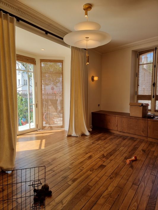 Un salon lumineux et vide, avec parquet, un luminaire suspendu à plusieurs niveaux, des rideaux beiges et un meuble bas en bois.