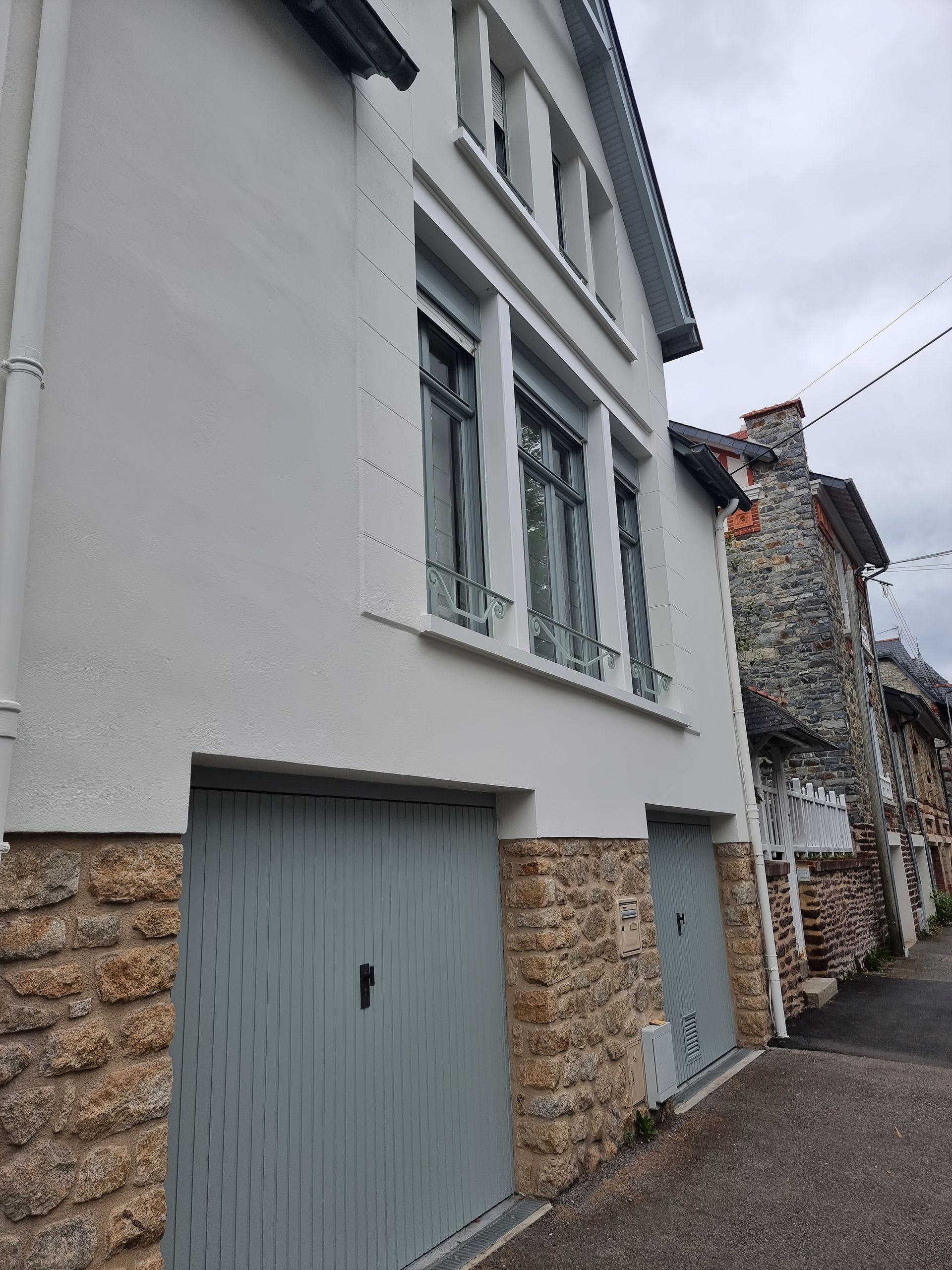 Une maison blanche avec un soubassement en pierre, deux portes de garage en bois gris et de hautes fenêtres à cadre gris, donnant sur une rue pavée.