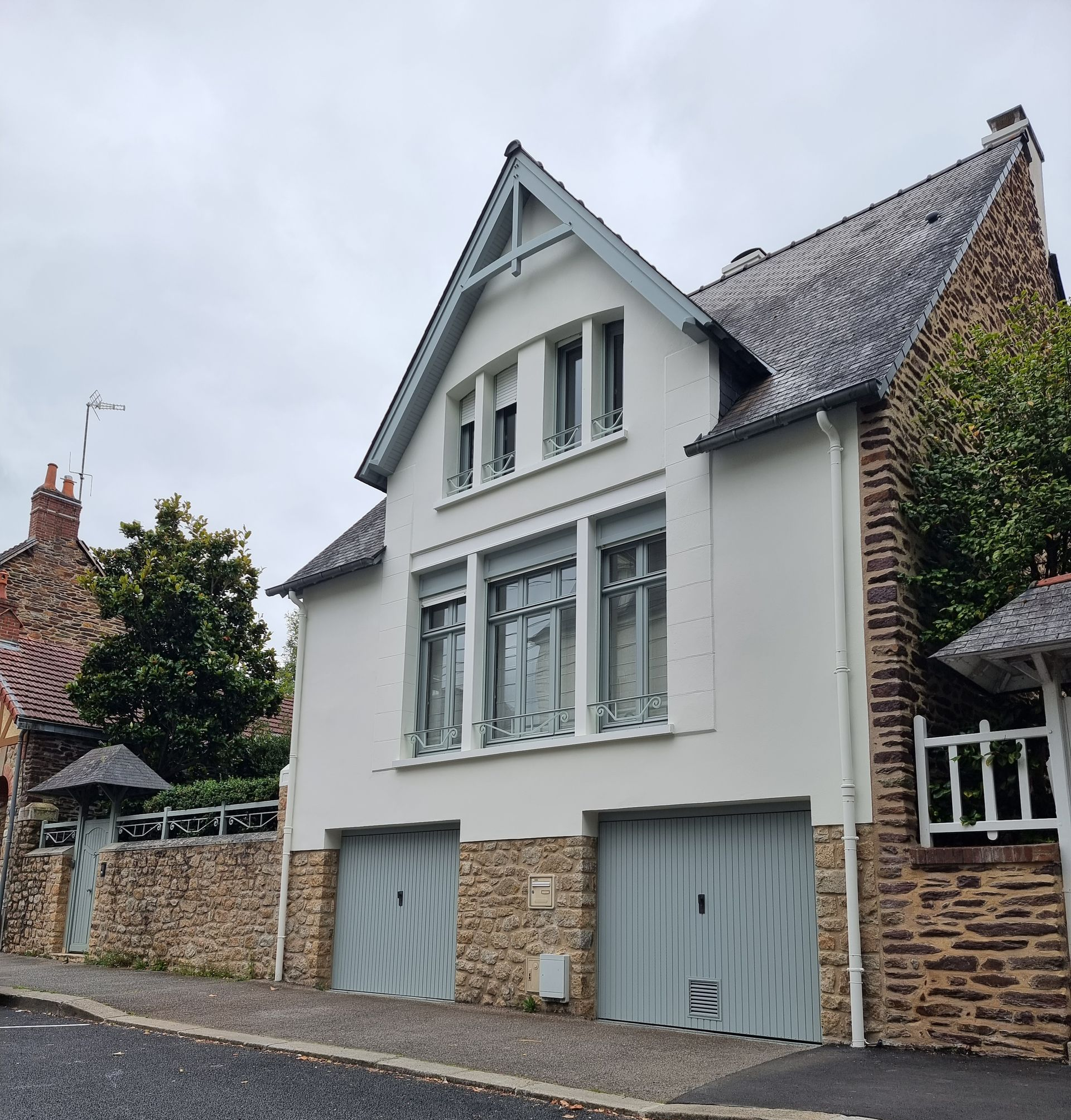 Une maison blanche à deux étages avec deux portes de garage bleu clair, des fondations en pierre et un toit sombre et pentu, située dans une rue pavée.