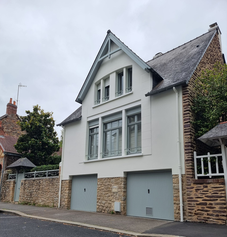 Une maison blanche à deux étages avec deux portes de garage bleu clair, des fondations en pierre et un toit sombre et pentu, située dans une rue pavée.