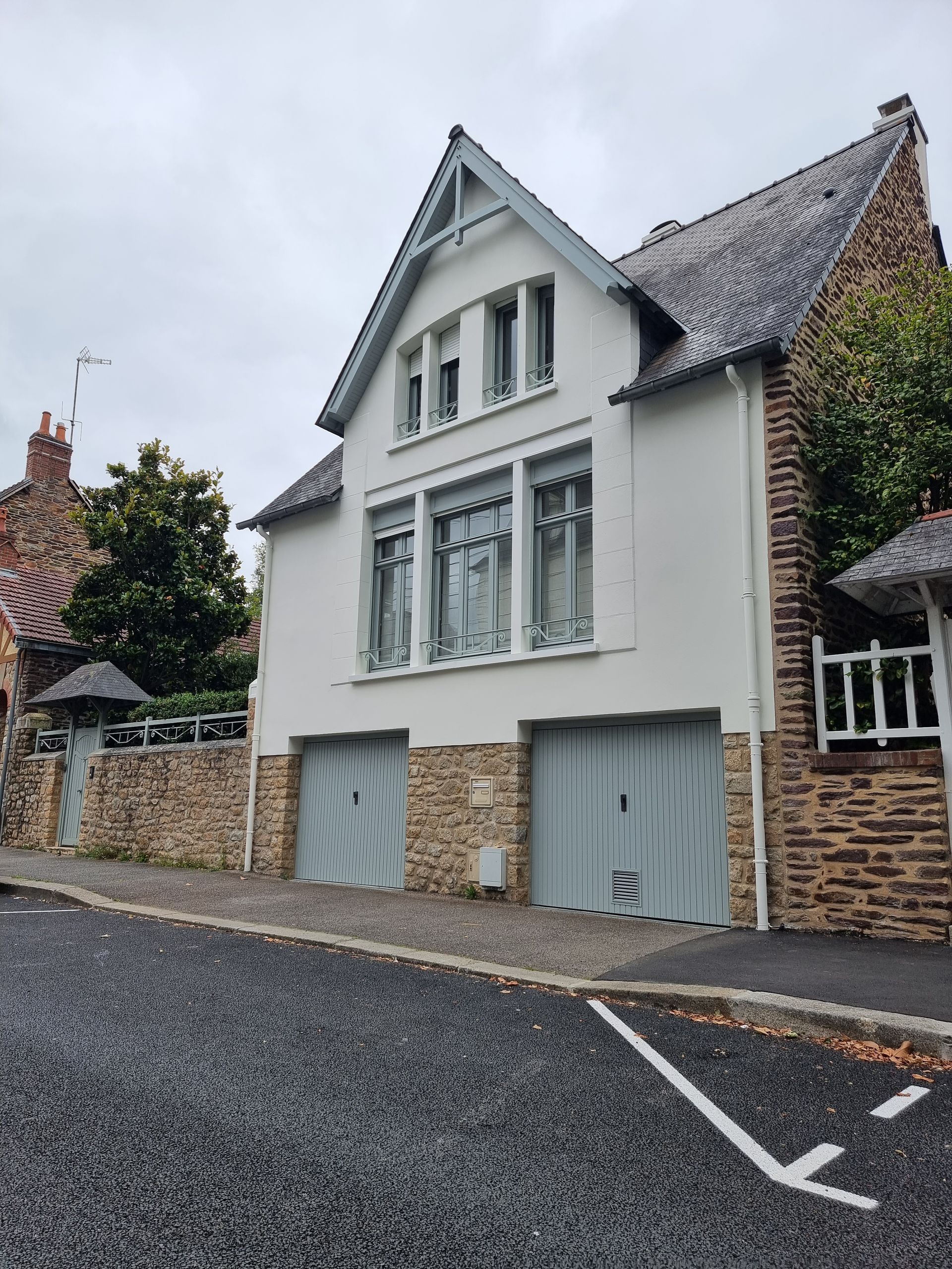 Une maison à plusieurs étages avec une façade blanche, un soubassement en pierre et deux portes de garage, située à côté d'un mur de pierre et d'un chemin de gravier.