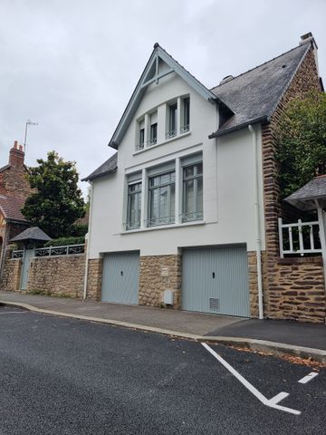 Une maison à plusieurs étages avec une façade blanche, un soubassement en pierre et deux portes de garage, située à côté d'un mur de pierre et d'un chemin de gravier.