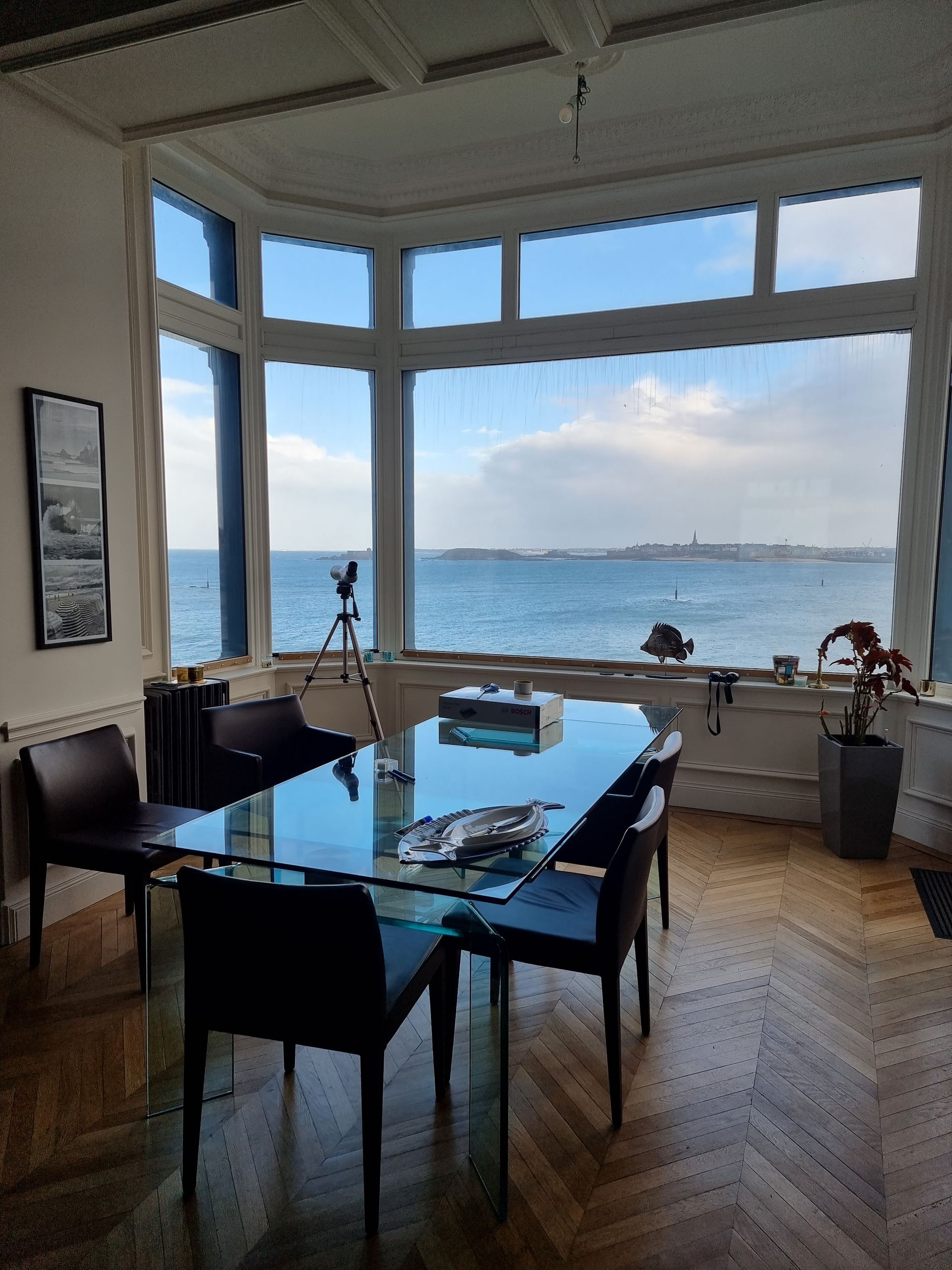Une table à manger avec un plateau en verre est placée devant une grande baie vitrée donnant sur une vue imprenable sur l'océan.