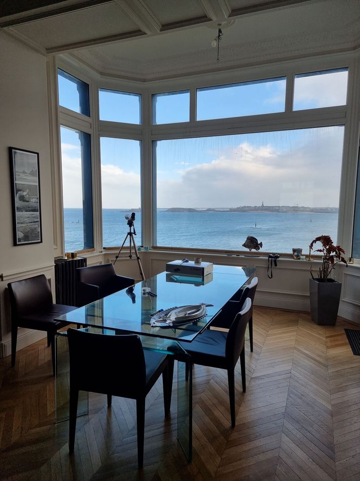 Une table à manger avec un plateau en verre est placée devant une grande baie vitrée donnant sur une vue imprenable sur l'océan.