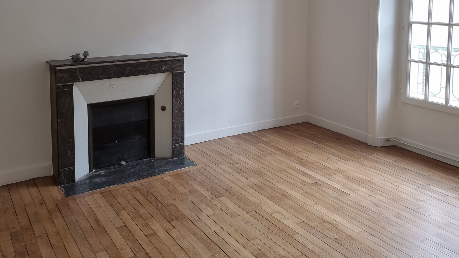 Une pièce vide avec un parquet clair, des murs blancs et une cheminée décorative en marbre foncé.