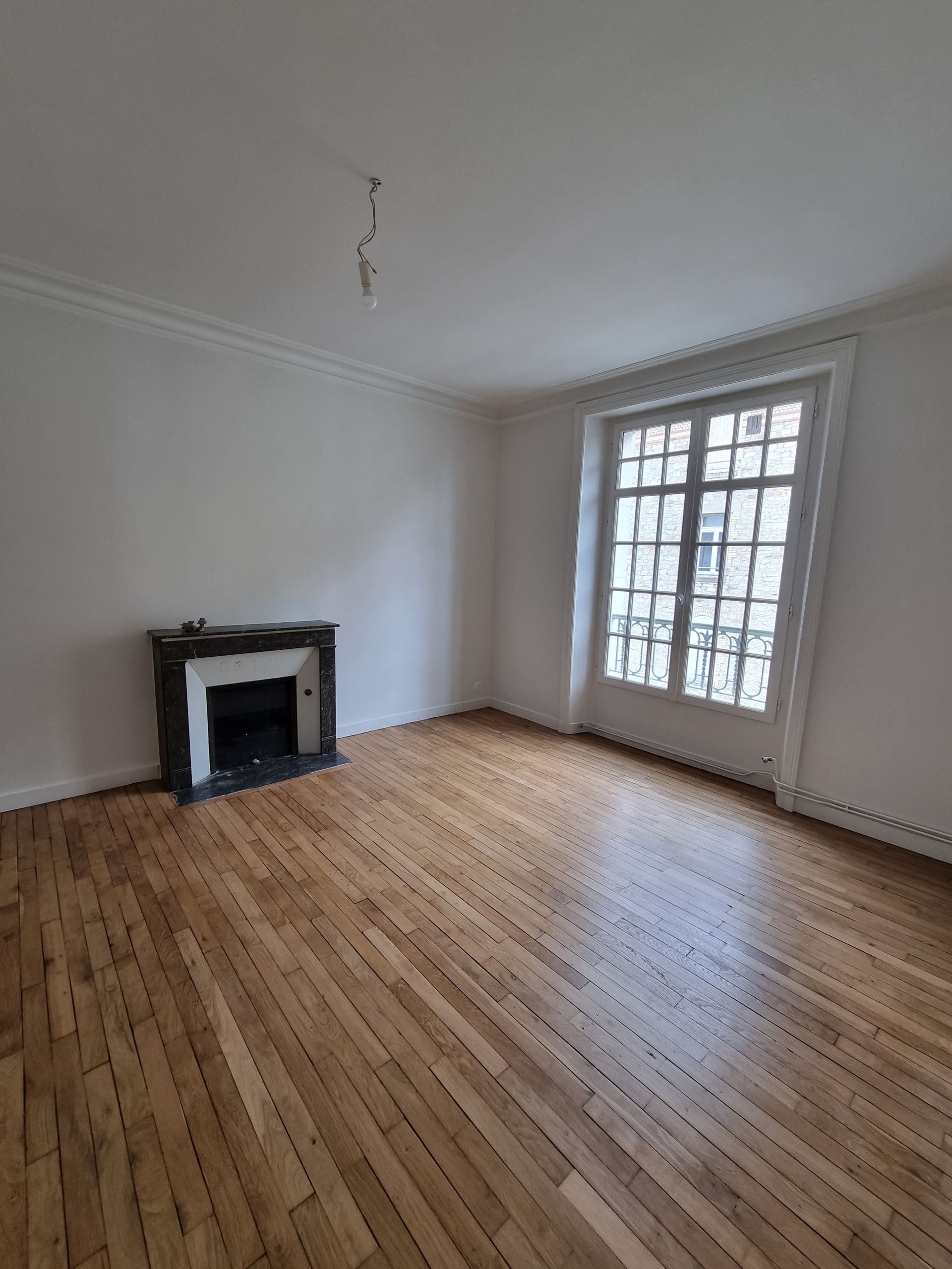 Une pièce vide aux murs blancs, avec une cheminée, un parquet et une grande fenêtre à motif quadrillé.