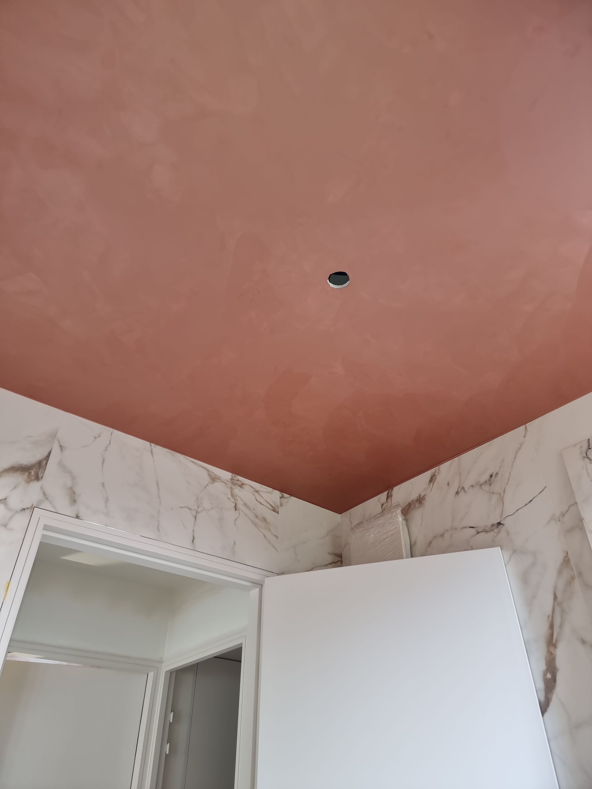 Un plafond lisse, couleur terre cuite, percé d'un trou central pour un luminaire, au-dessus de murs revêtus de carreaux blancs imitation marbre.