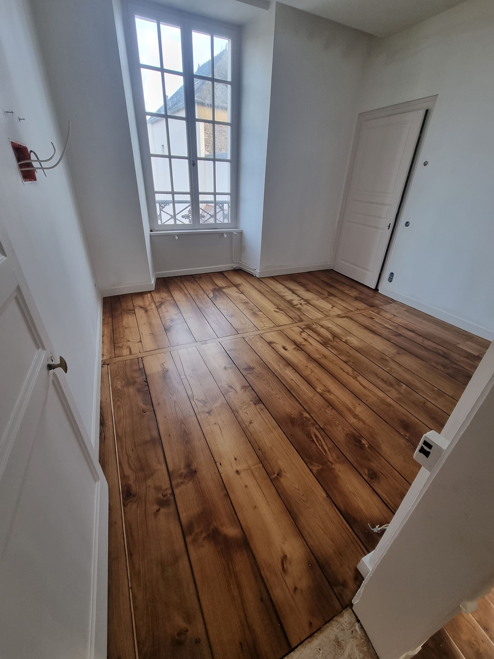 Une pièce vide aux murs clairs, avec une grande fenêtre, des portes blanches et un parquet en bois naturel à larges lames.