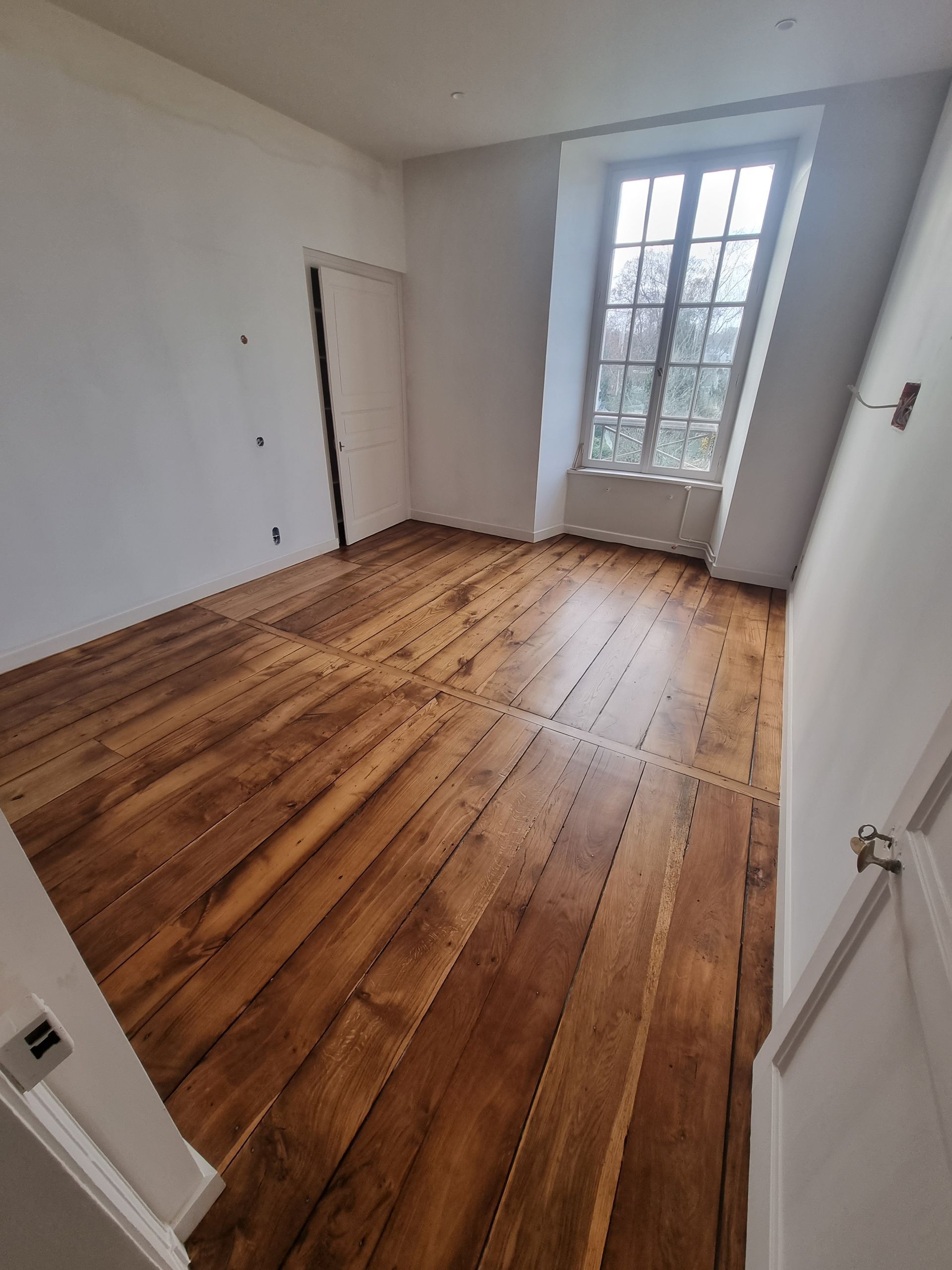 Une pièce vide avec un parquet à larges lattes, des murs blancs et une haute fenêtre donnant sur l'extérieur.