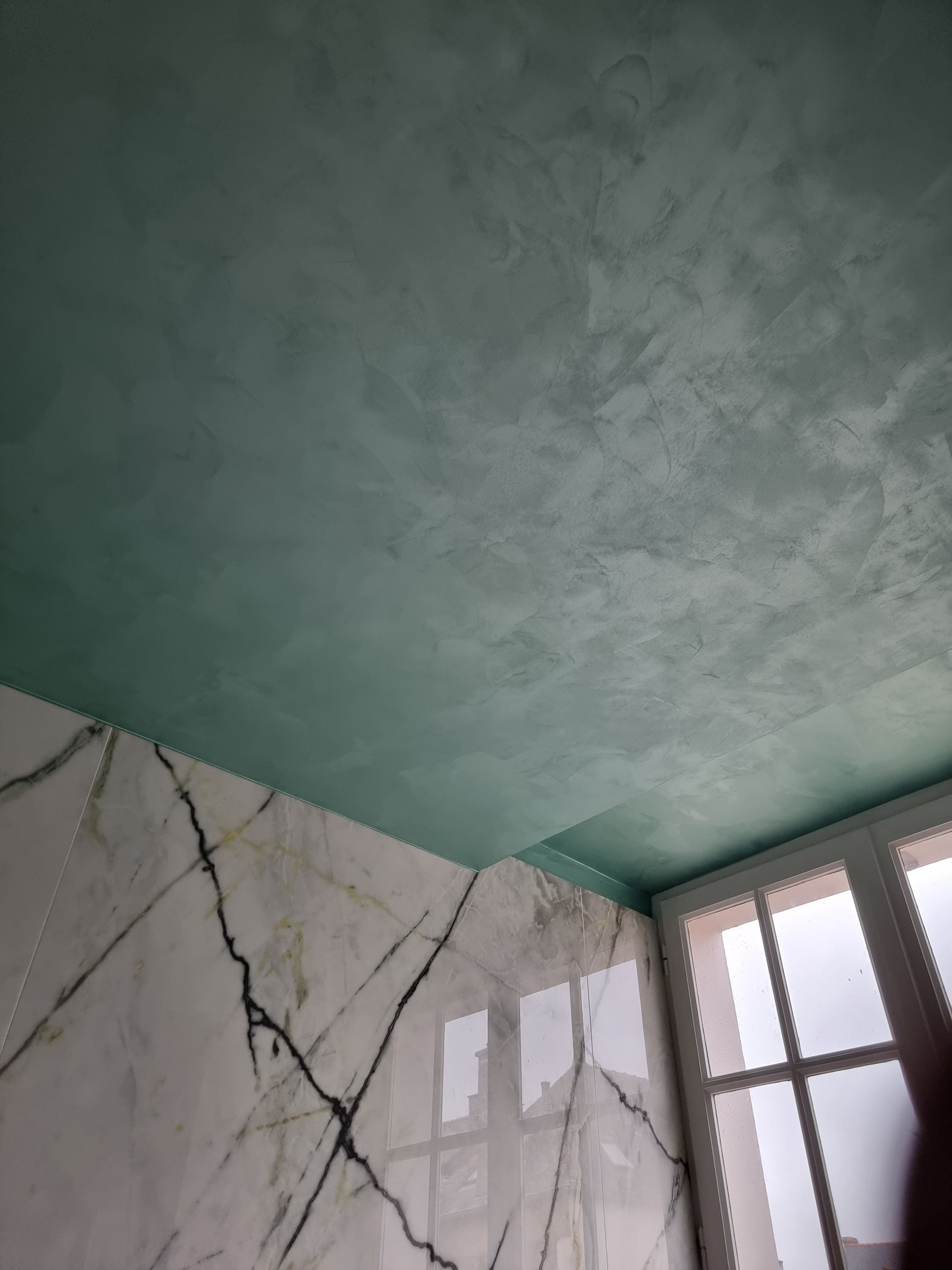Un plafond peint en vert turquoise texturé contraste avec un mur en marbre blanc veiné de noir, près d'une fenêtre.