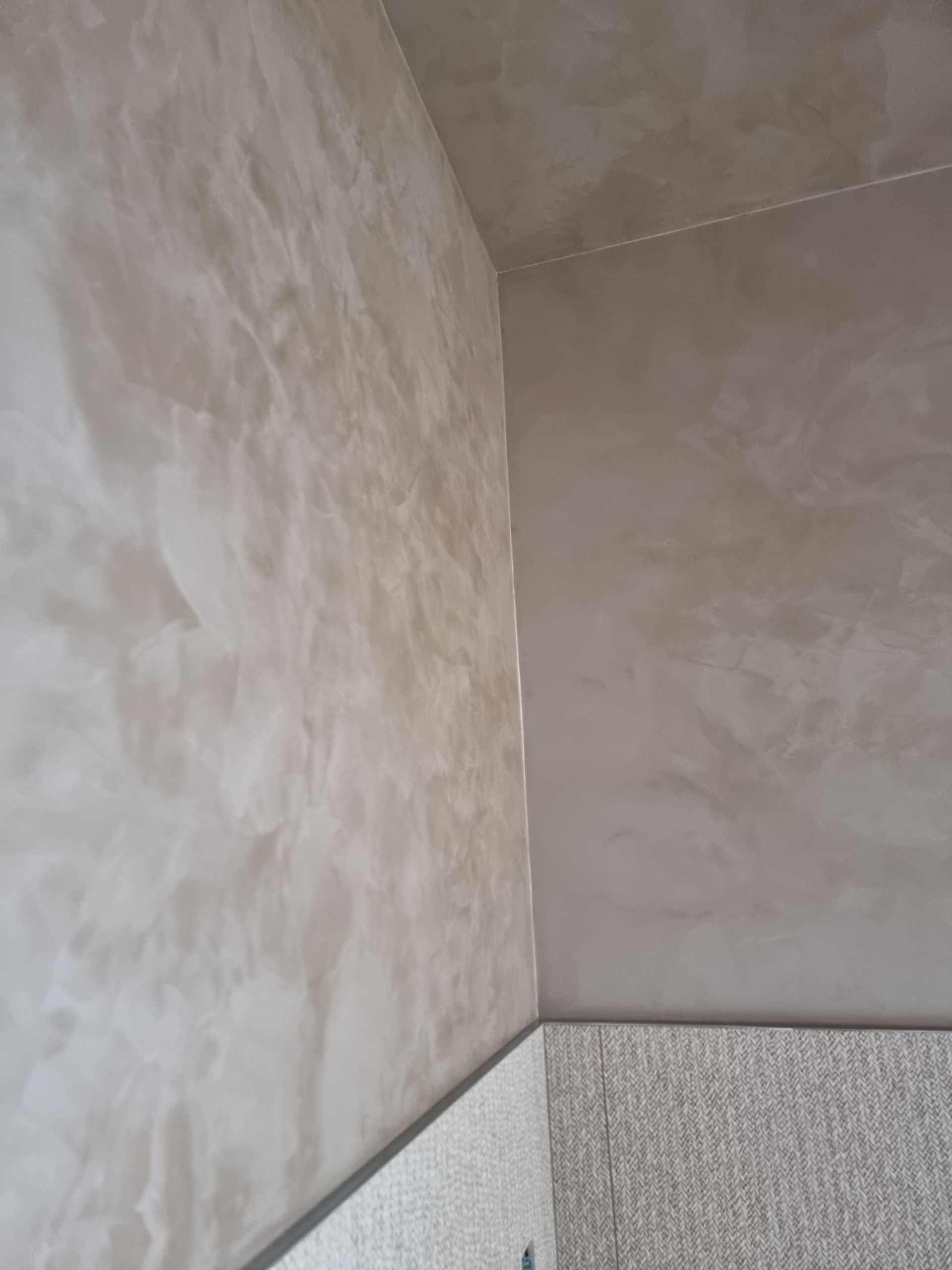 Un angle intérieur où un mur texturé beige rencontre un mur plat aux tons gris au-dessus de sections carrelées de couleur claire.