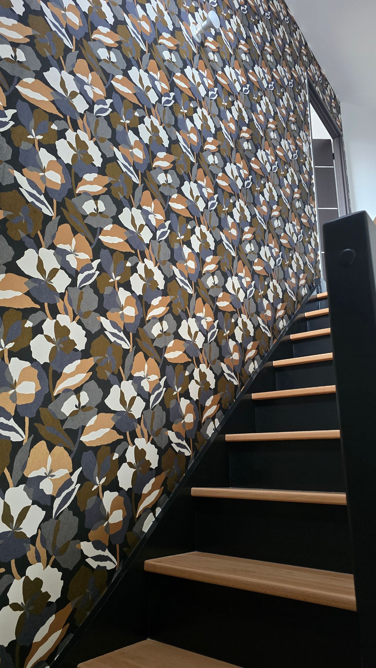 Escalier avec contremarches noires, marches en bois et rampe noire, adossé à un mur recouvert d'un papier peint à motifs floraux foncés.