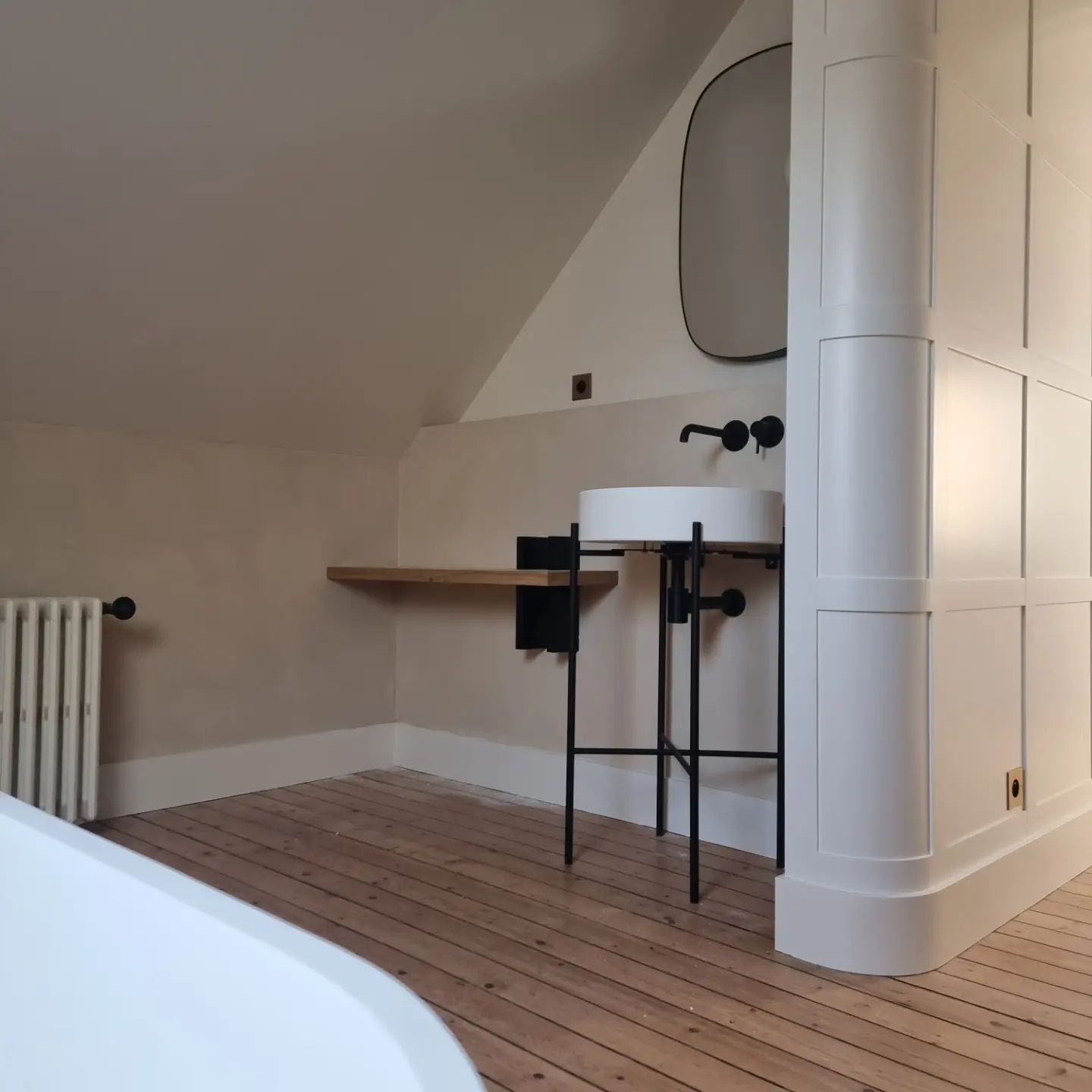Une salle de bains minimaliste avec un lavabo sur pied, un miroir arqué, un parquet et une cloison murale incurvée à panneaux.