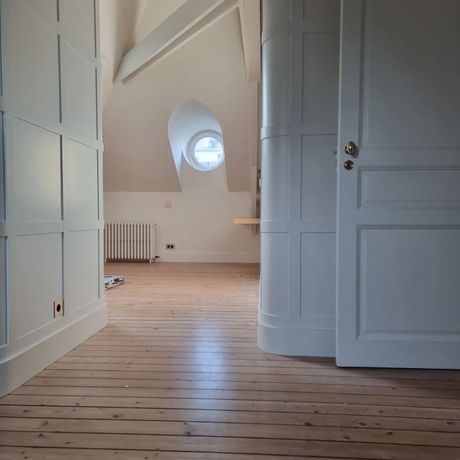 Un couloir mansardé avec un parquet clair, des murs lambrissés incurvés, une porte unique et une fenêtre circulaire au loin.