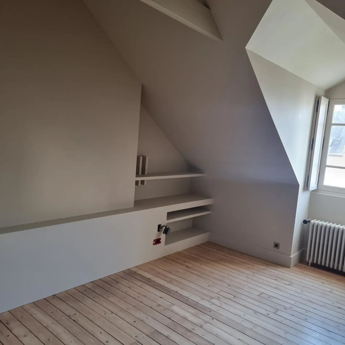 Une pièce mansardée vide, avec des murs clairs, un plafond mansardé, un parquet, des étagères intégrées et une fenêtre avec un radiateur.