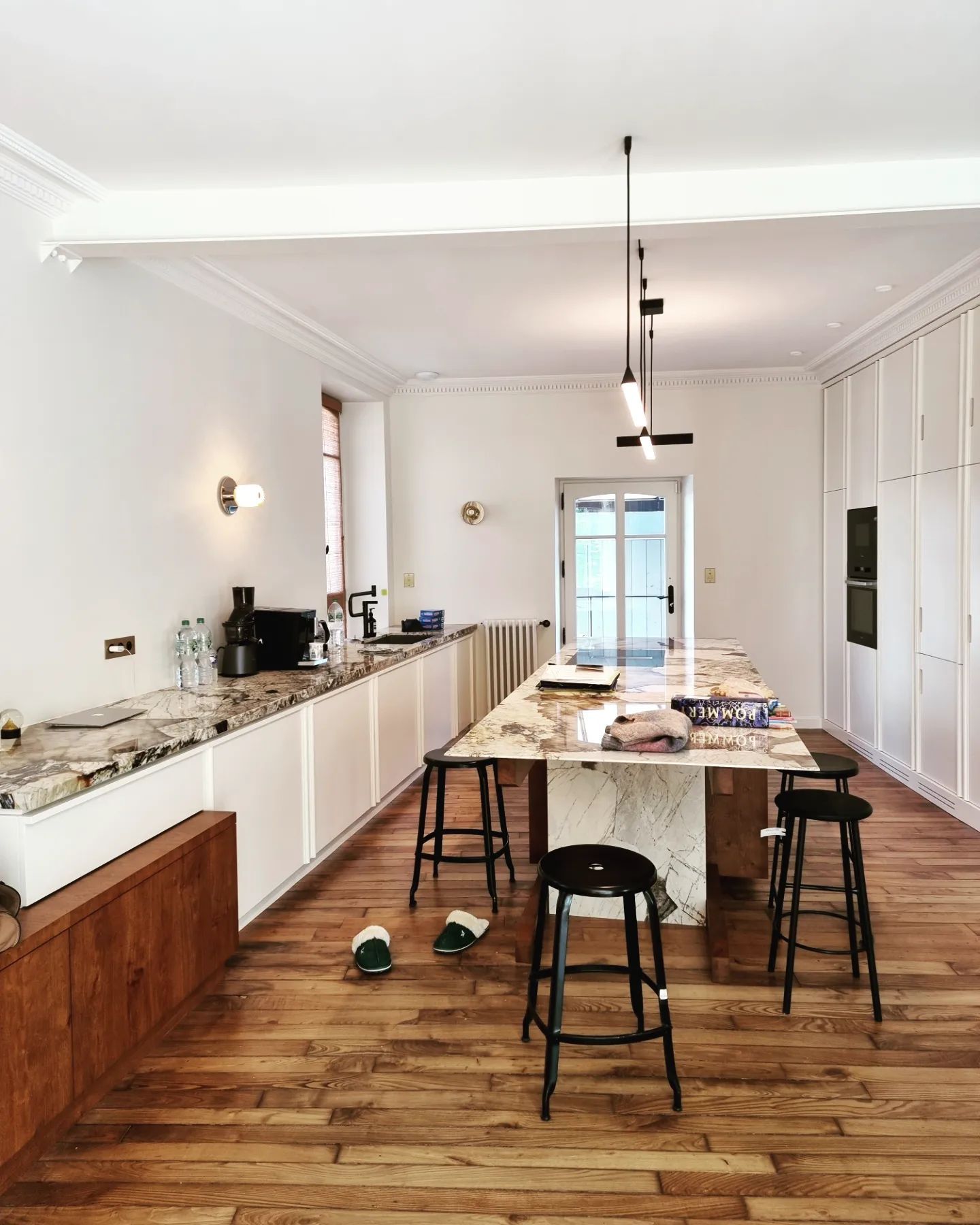 Une cuisine moderne avec des armoires blanches, un îlot central en marbre, des tabourets noirs et un parquet sous un haut plafond.