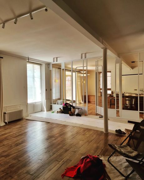 Une pièce lumineuse et aérée avec parquet, une cloison en métal blanc et un morceau de tissu rouge au premier plan.