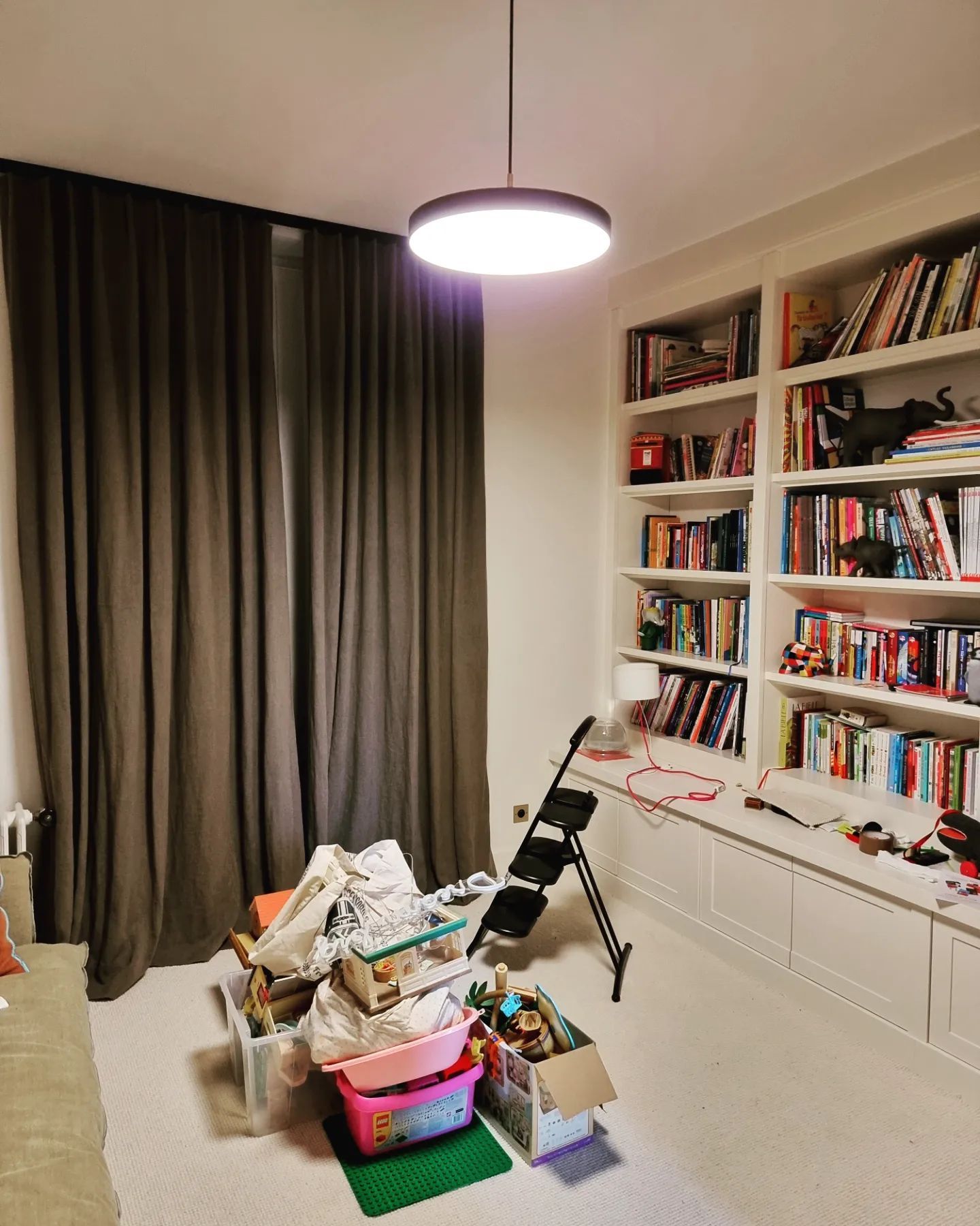 Une pièce avec une bibliothèque, un rideau gris, une suspension lumineuse et un sol encombré d'objets, dont des poubelles et un petit escabeau.