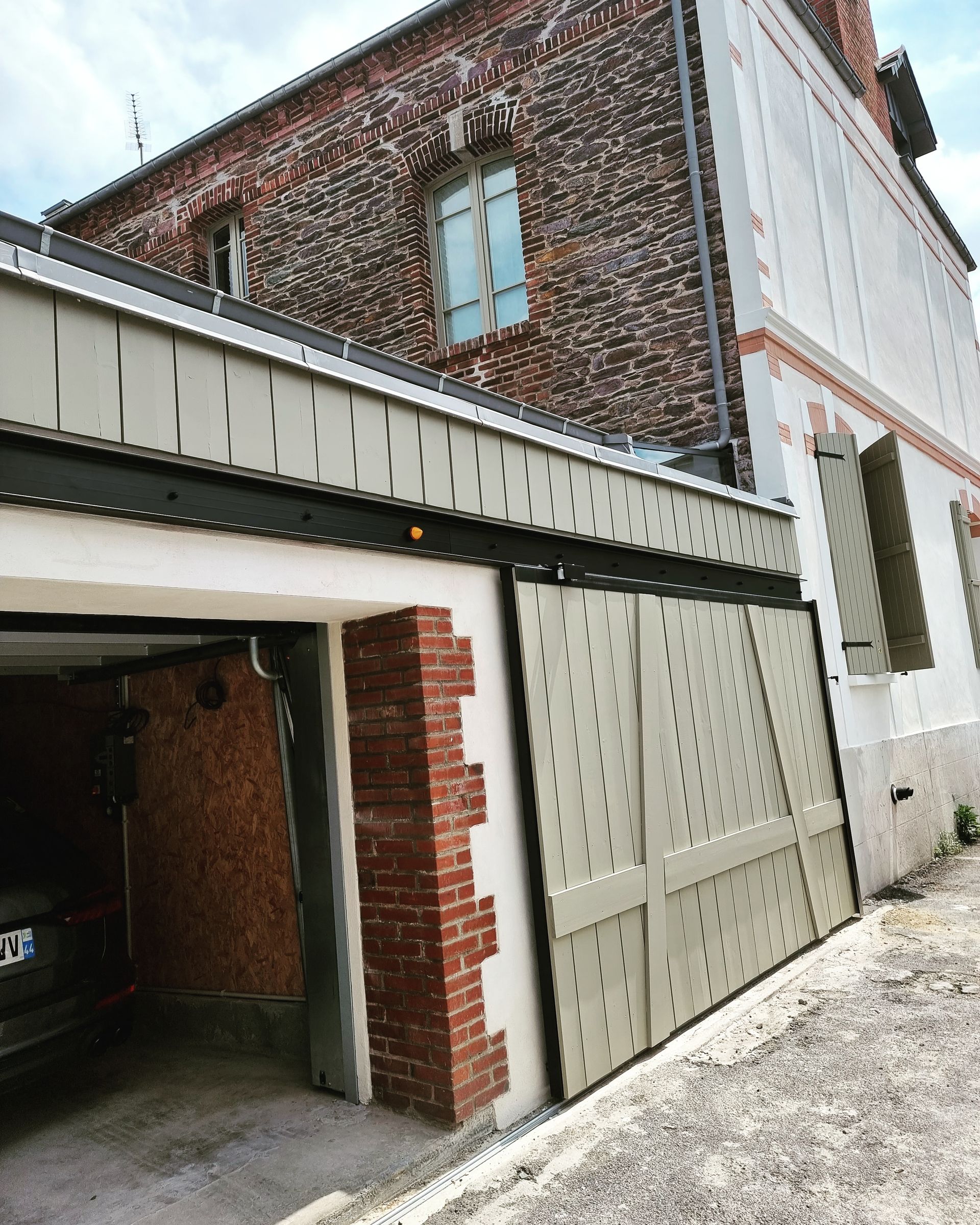 Une porte de garage coulissante en métal perforé, fixée à une maison historique en briques avec des volets extérieurs.