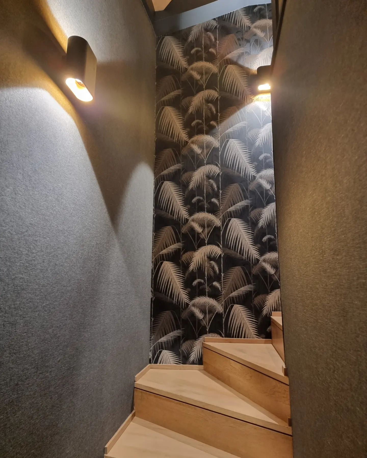 Un escalier en bois aux marches claires, flanqué de murs à la texture sombre et éclairé par deux appliques murales à la lumière chaude.