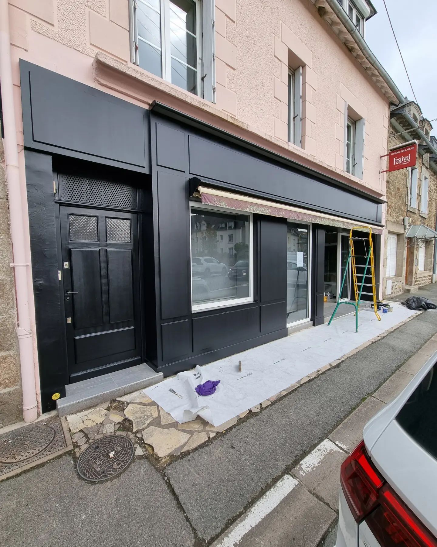 Une devanture de magasin avec une façade noire fraîchement peinte, de grandes fenêtres, une porte noire et une échelle visible à l'intérieur.