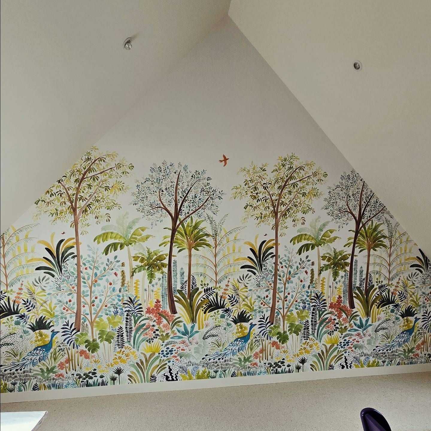 Un papier peint aux couleurs vives, sur le thème de la jungle, avec des arbres colorés, des plantes exotiques et un paon sur un mur de grenier en pente.