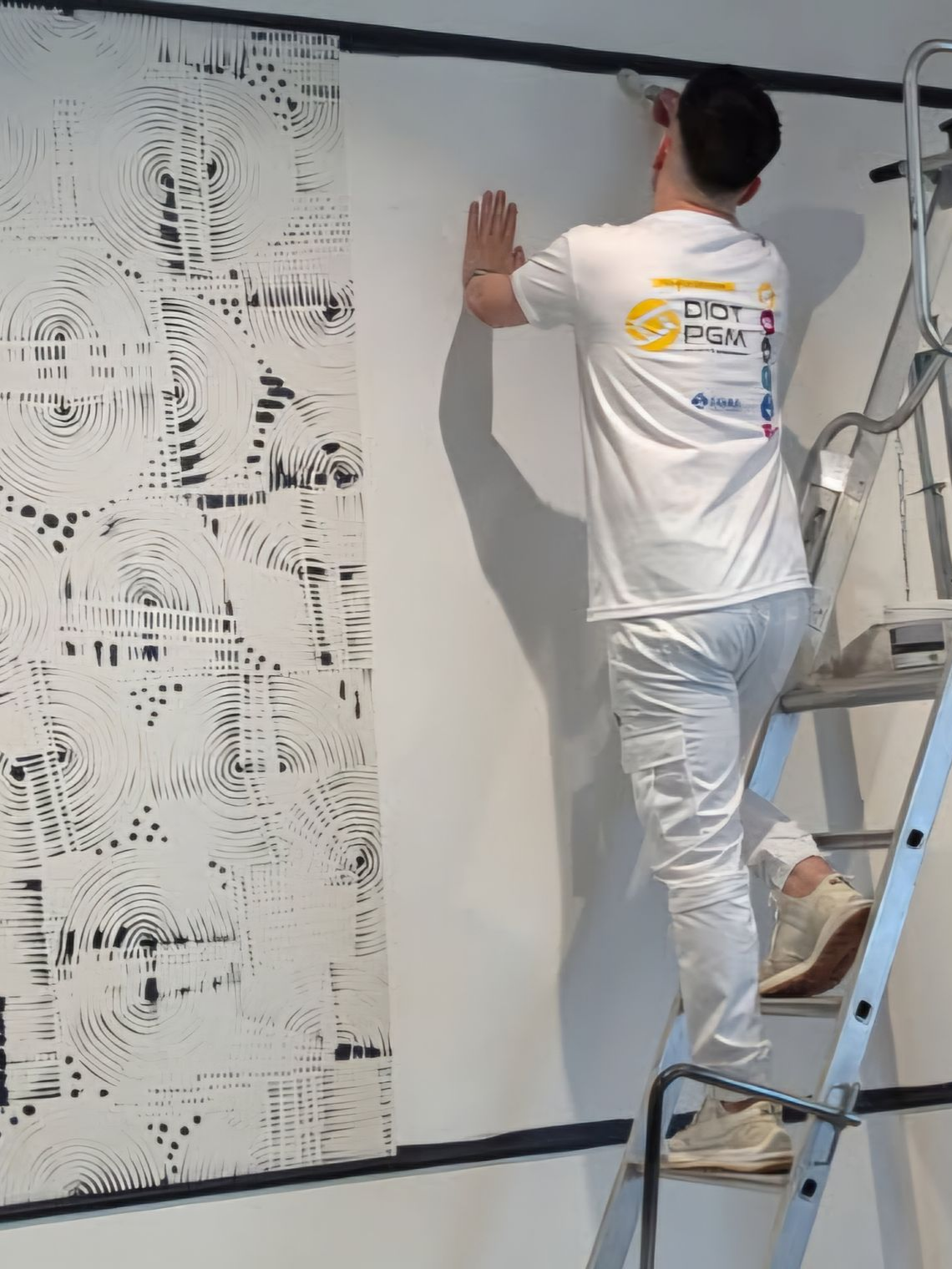 Une personne sur une échelle peint une ligne noire horizontale sur un mur blanc, à côté d'une estampe abstraite verticale en noir et blanc.